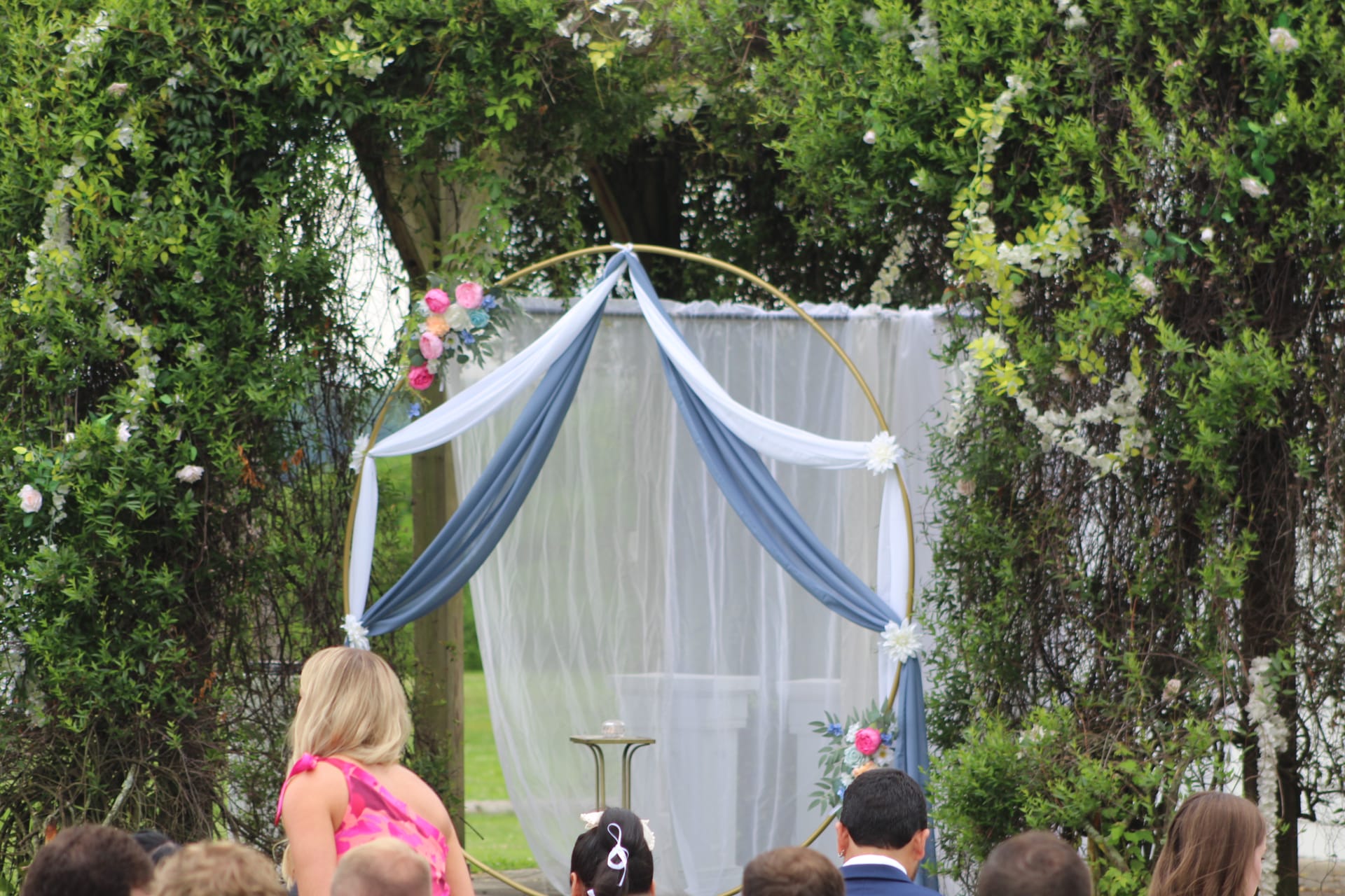 A gold circular hoop wedding arch draped in dusty blue and white fabric with pink and white floral accents stands beneath lush green trees at an outdoor garden ceremony at Scarborough House.