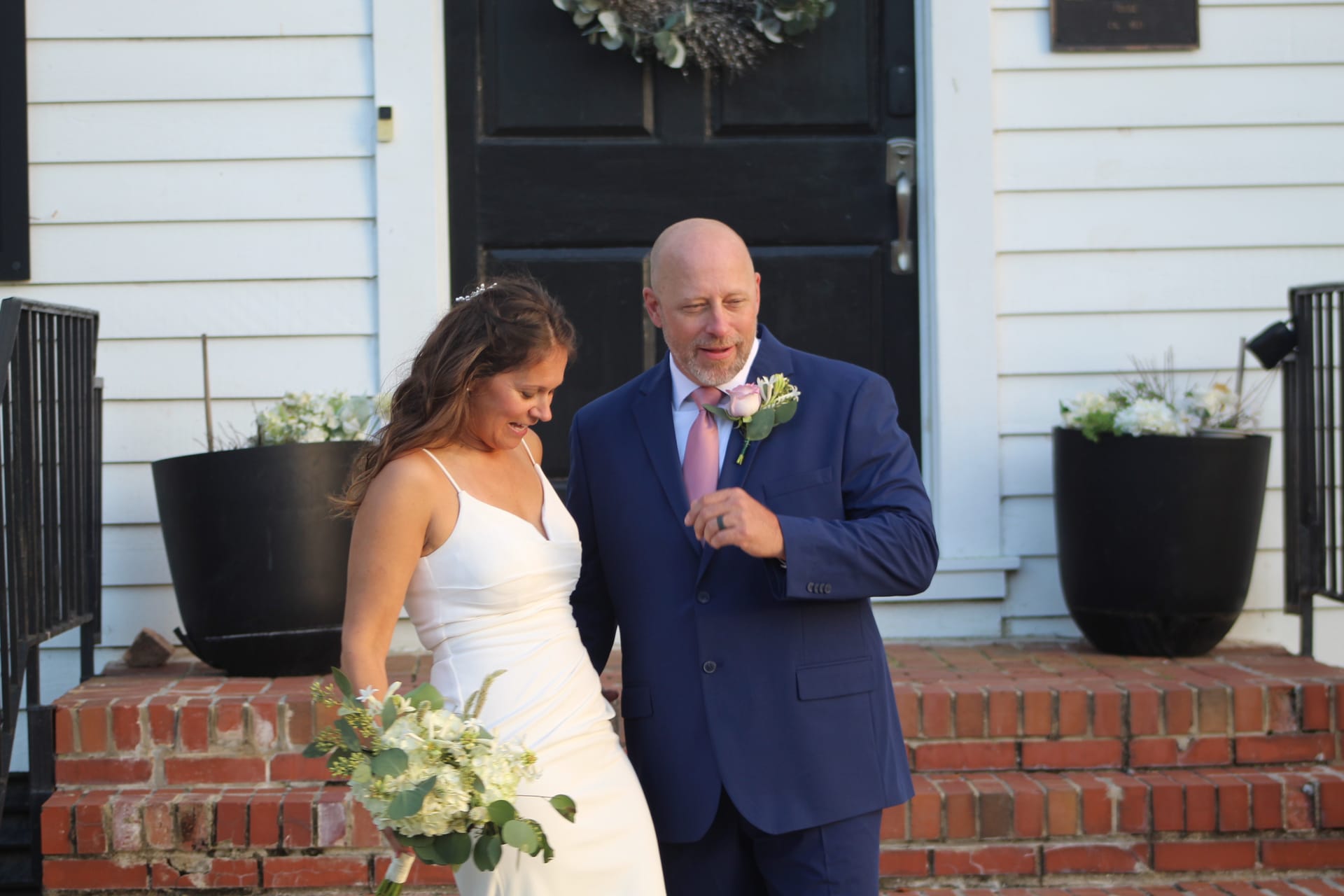 A bride in a white gown holding a lush white bouquet walks down the brick front steps of Scarborough House alongside a man in a navy suit with a pink tie and boutonniere.