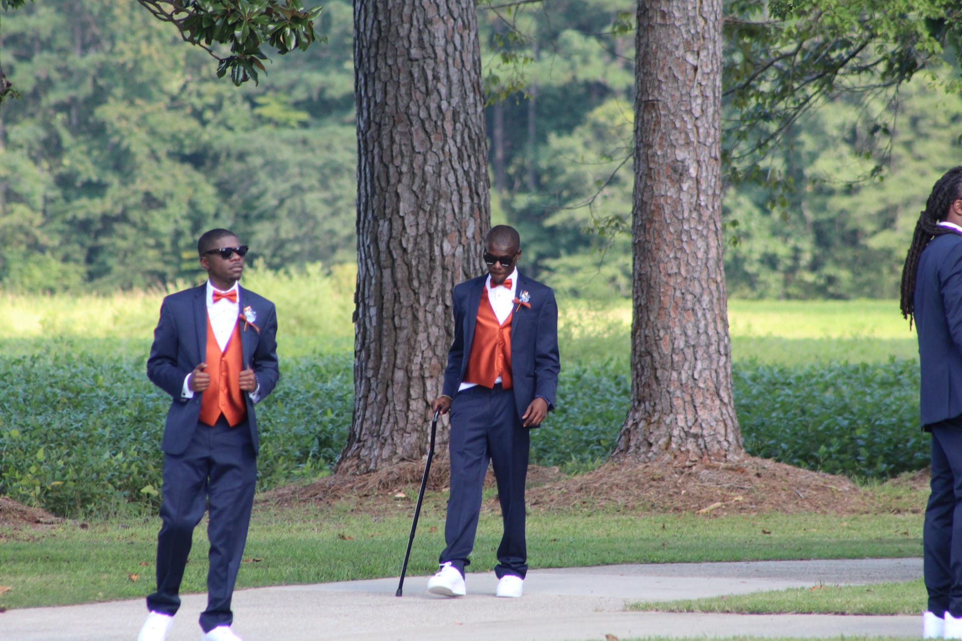 Three groomsmen in navy suits with burnt-orange vests and bow ties stand among large oak trees on the grounds of a historic estate wedding venue.