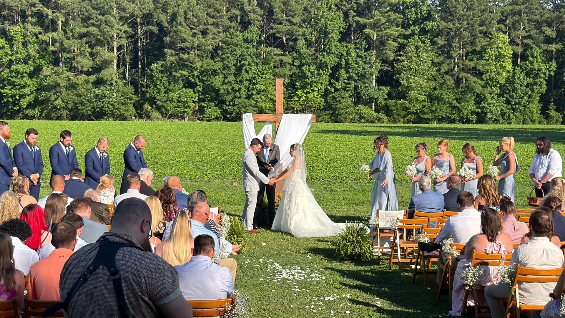 Bride and groom exchange vows beneath a draped wooden cross arch on the ceremony lawn at Scarborough House, flanked by groomsmen and bridesmaids in blue while guests look on from wooden chairs.