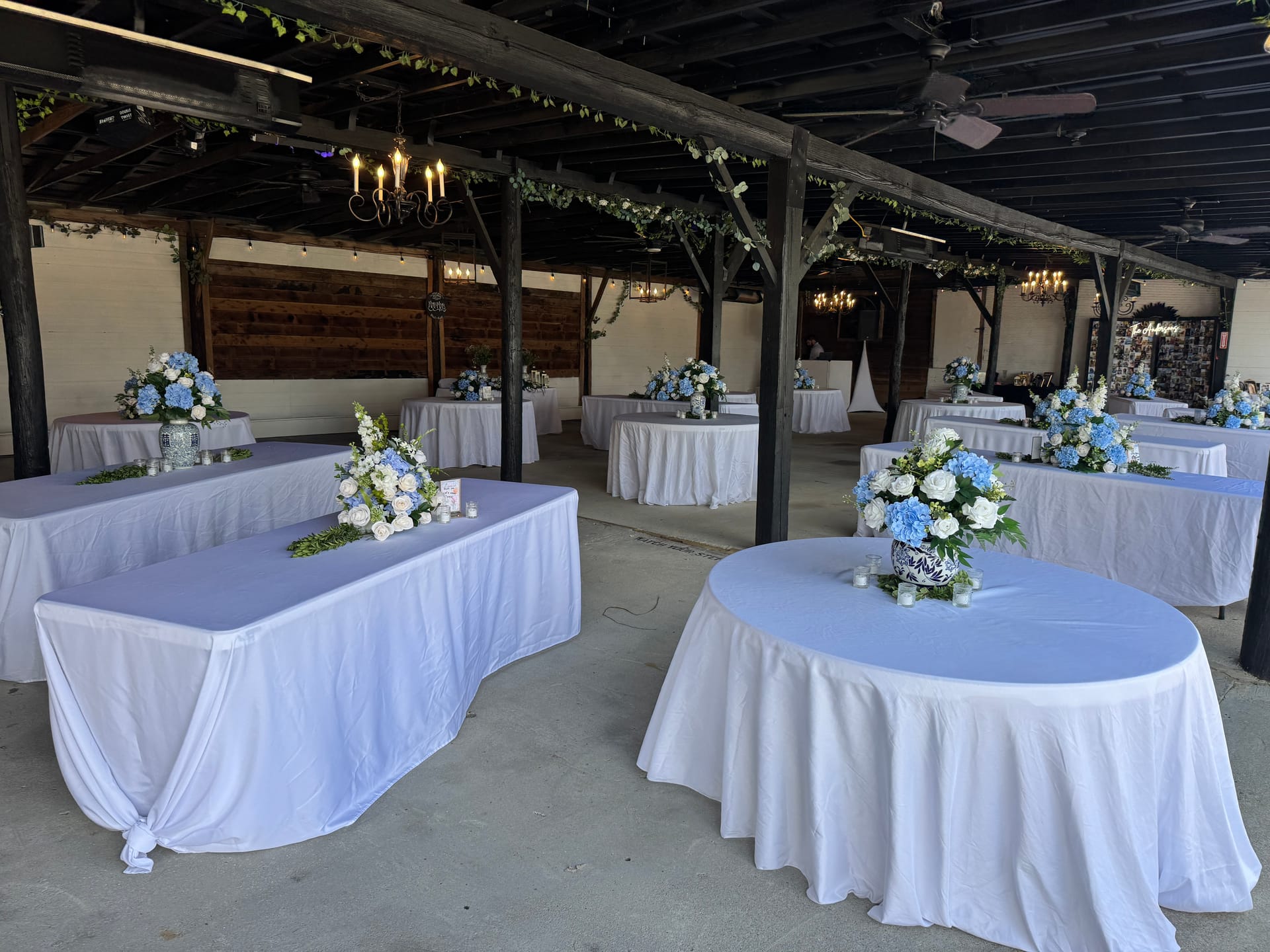 Elegantly decorated magnolia pavilion reception space with white-draped tables adorned with blue hydrangea and white rose centerpieces beneath rustic wooden beams strung with greenery and chandeliers.