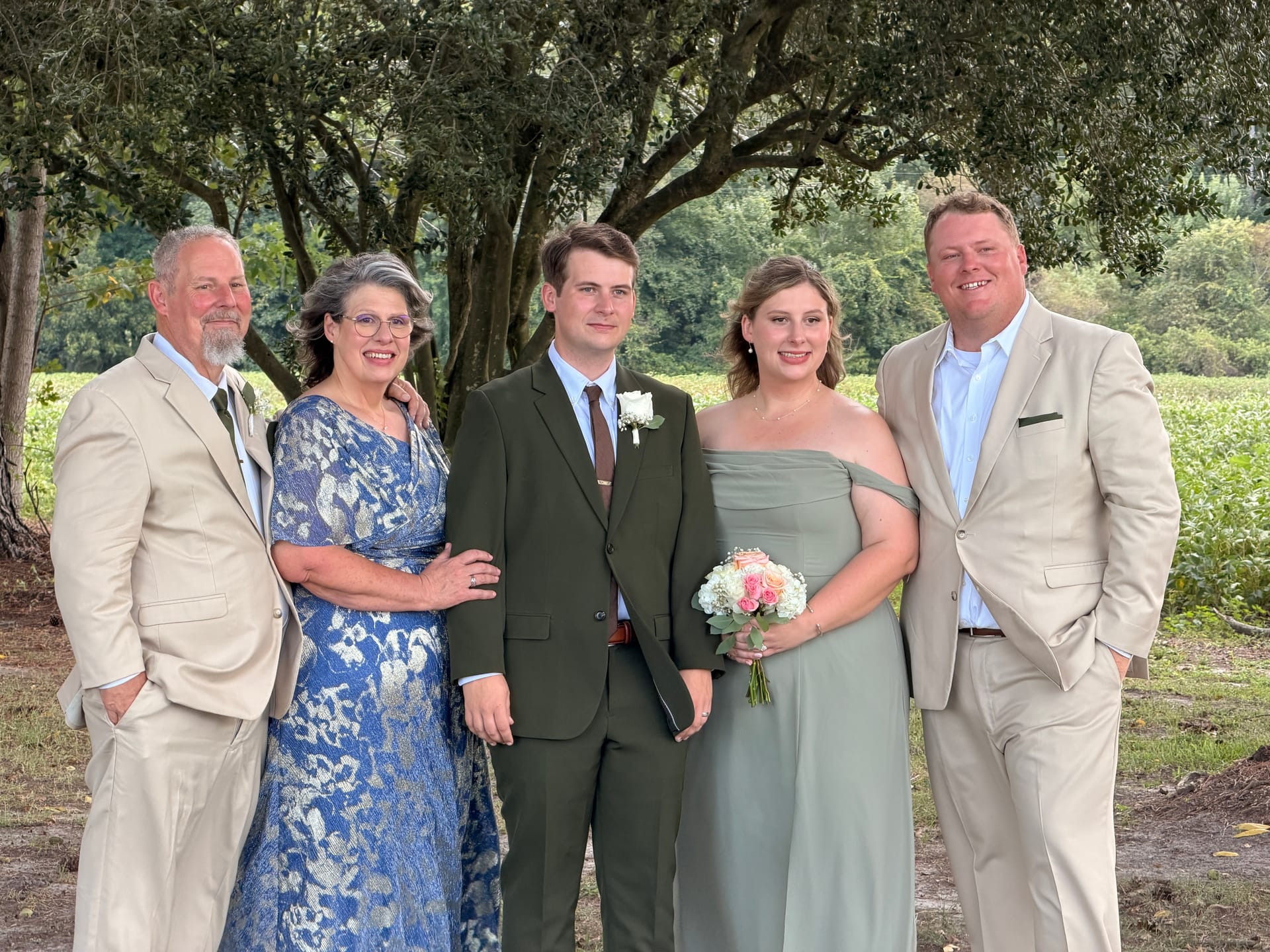 A family of five dressed in wedding attire smiles together beneath sprawling oak trees with green farmland fields in the background at Scarborough House estate.