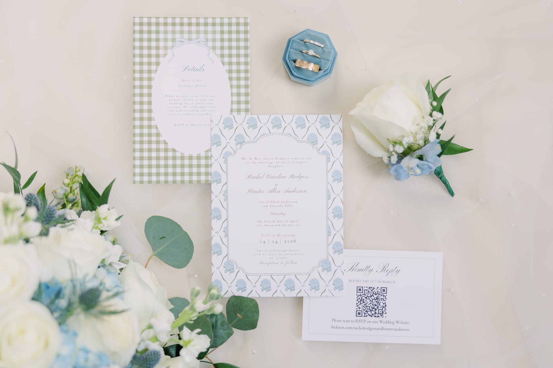 Flat lay of a blue and white floral invitation suite with gingham details, a QR code RSVP card, wedding rings in a velvet box, and a white rose boutonniere.