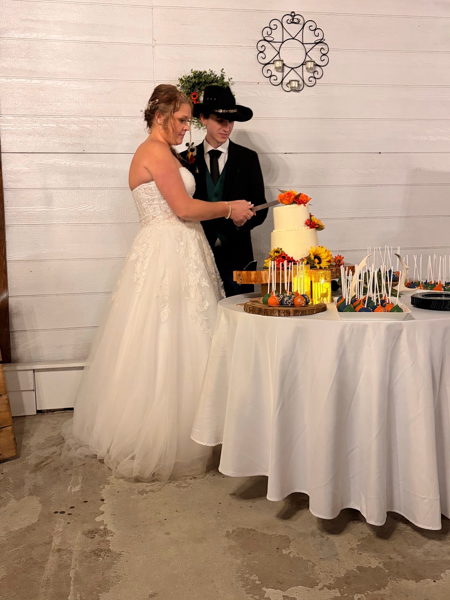 Bride in a white lace gown and groom in a black suit and cowboy hat cut their two-tier white wedding cake adorned with fall sunflowers and orange blooms at a rustic indoor reception.