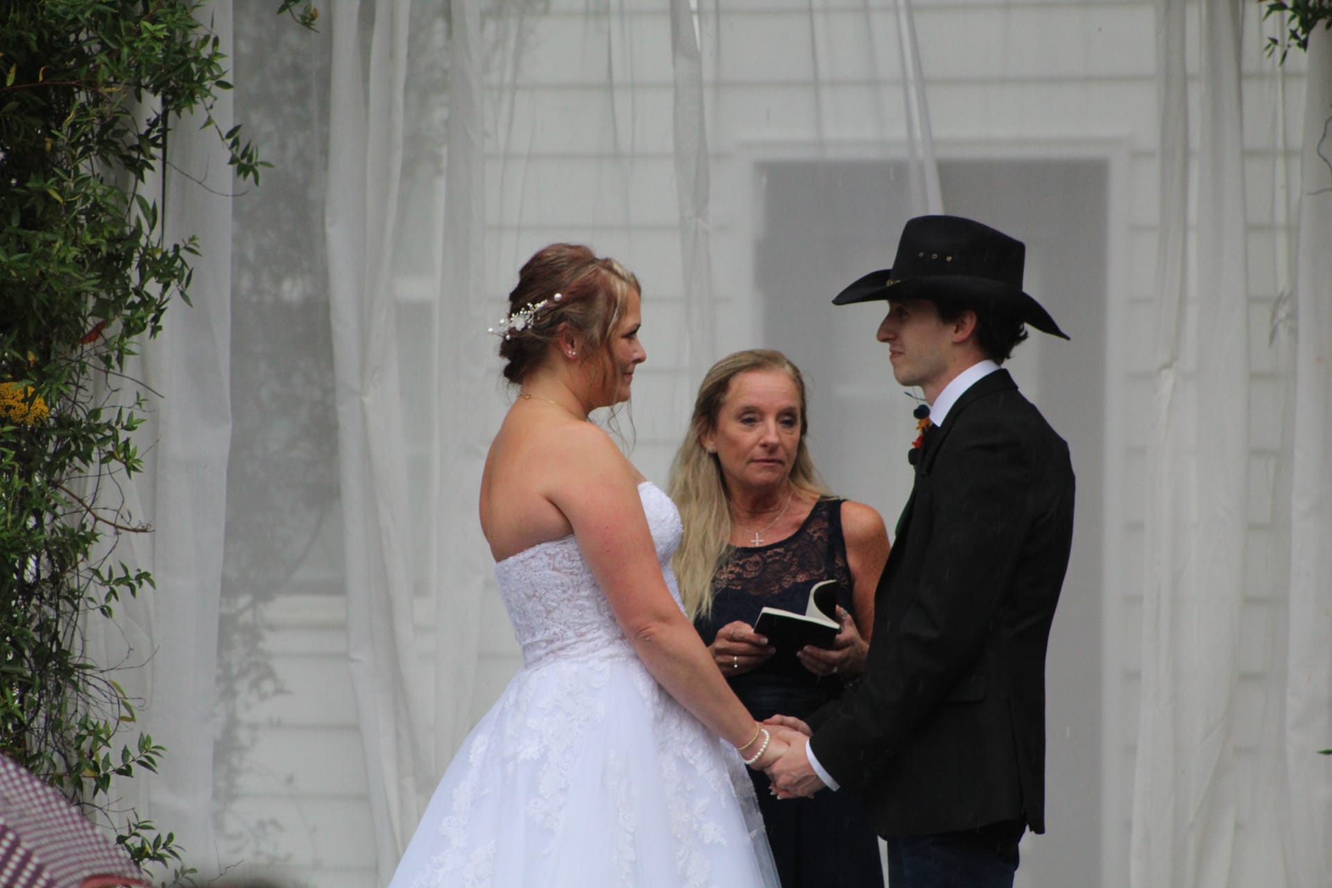 Bride in a white lace gown and groom in a black suit and cowboy hat exchange vows with an officiant holding a book during an outdoor ceremony at Scarborough House historic estate.