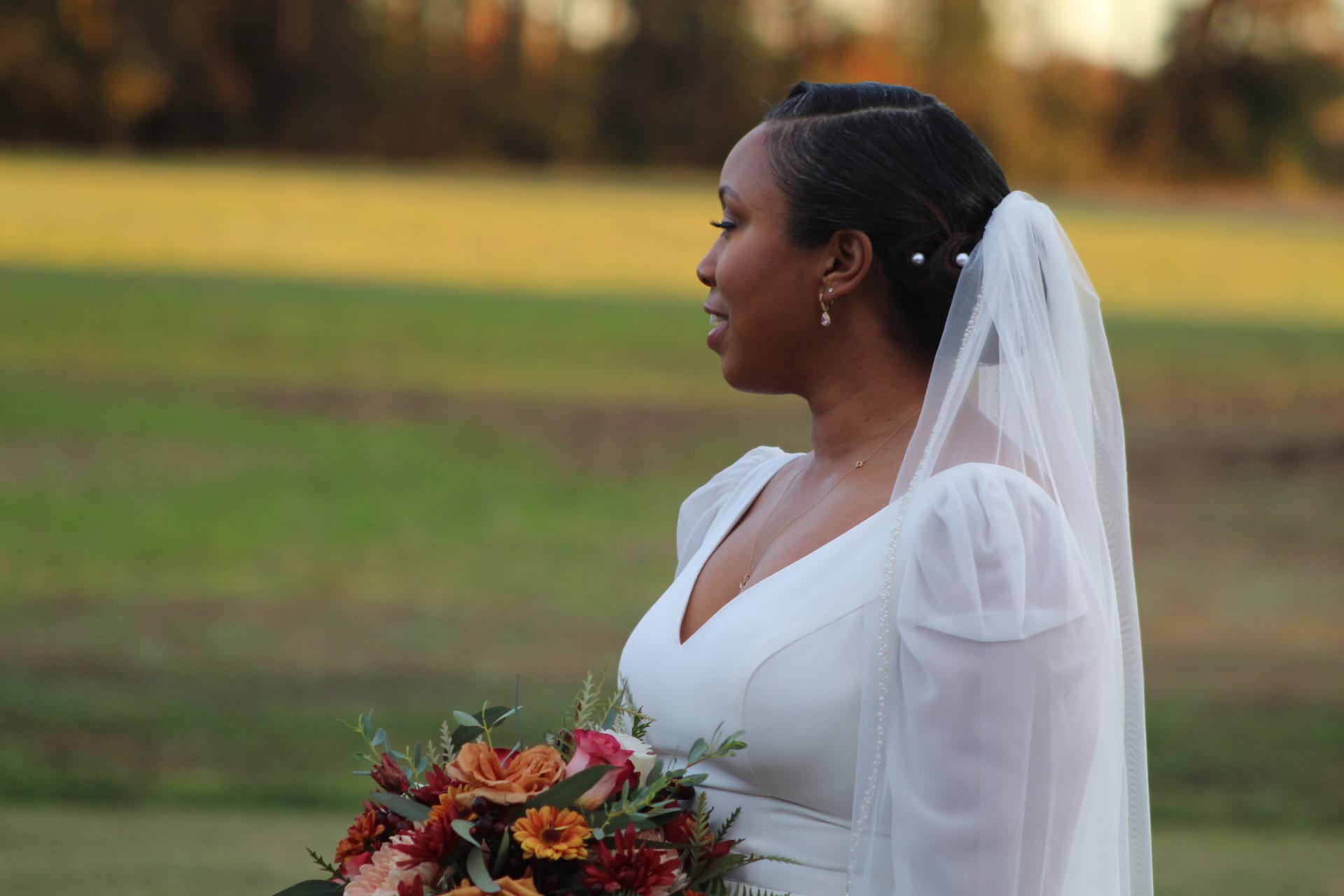 Bride in a white long-sleeve gown and pearl-trimmed veil holds a vibrant autumn bouquet of orange and burgundy flowers while gazing serenely across the sunlit fields at Scarborough House.