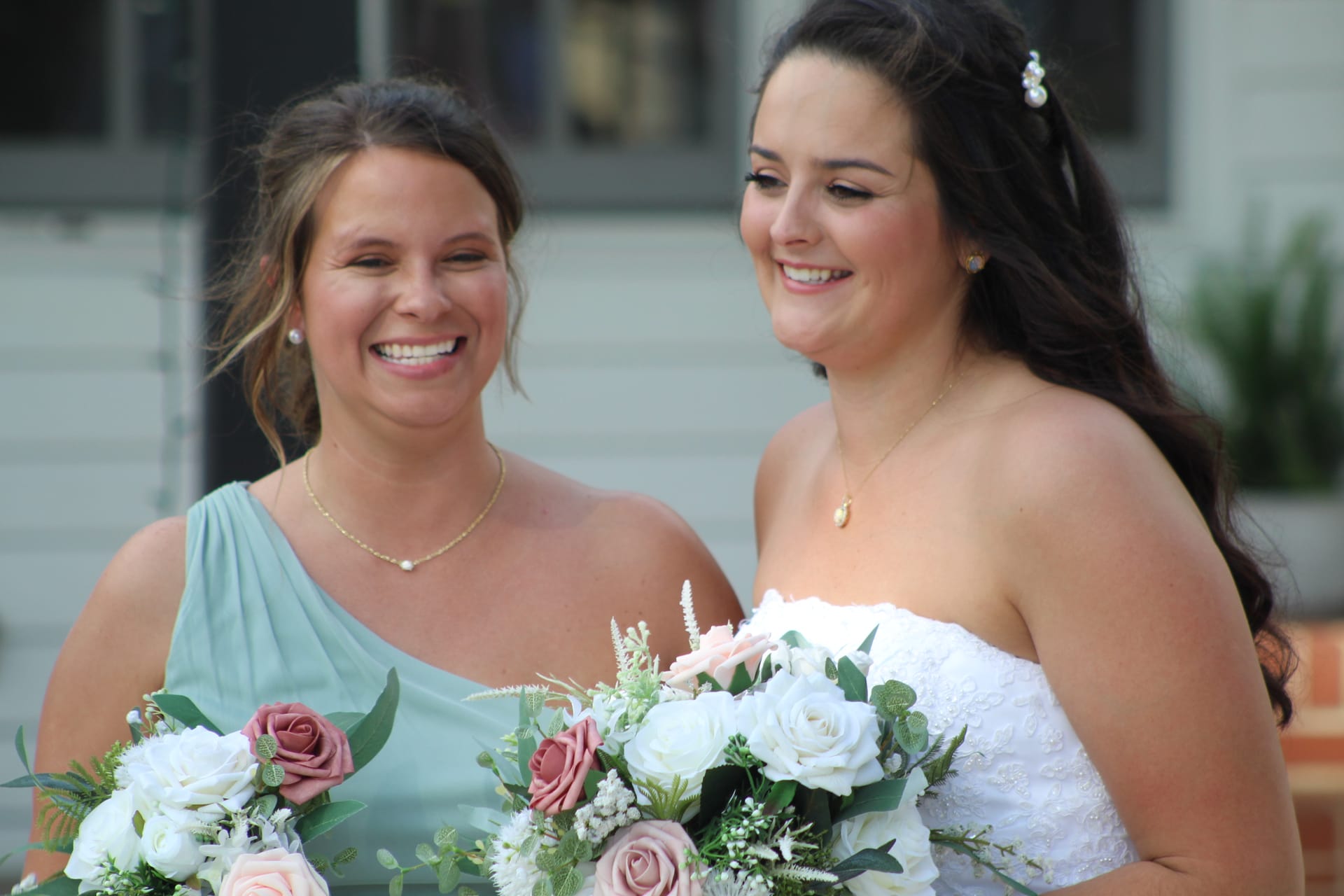 Smiling bride in a white strapless gown and her bridesmaid in sage green holding blush and white rose bouquets outside a historic estate wedding venue.