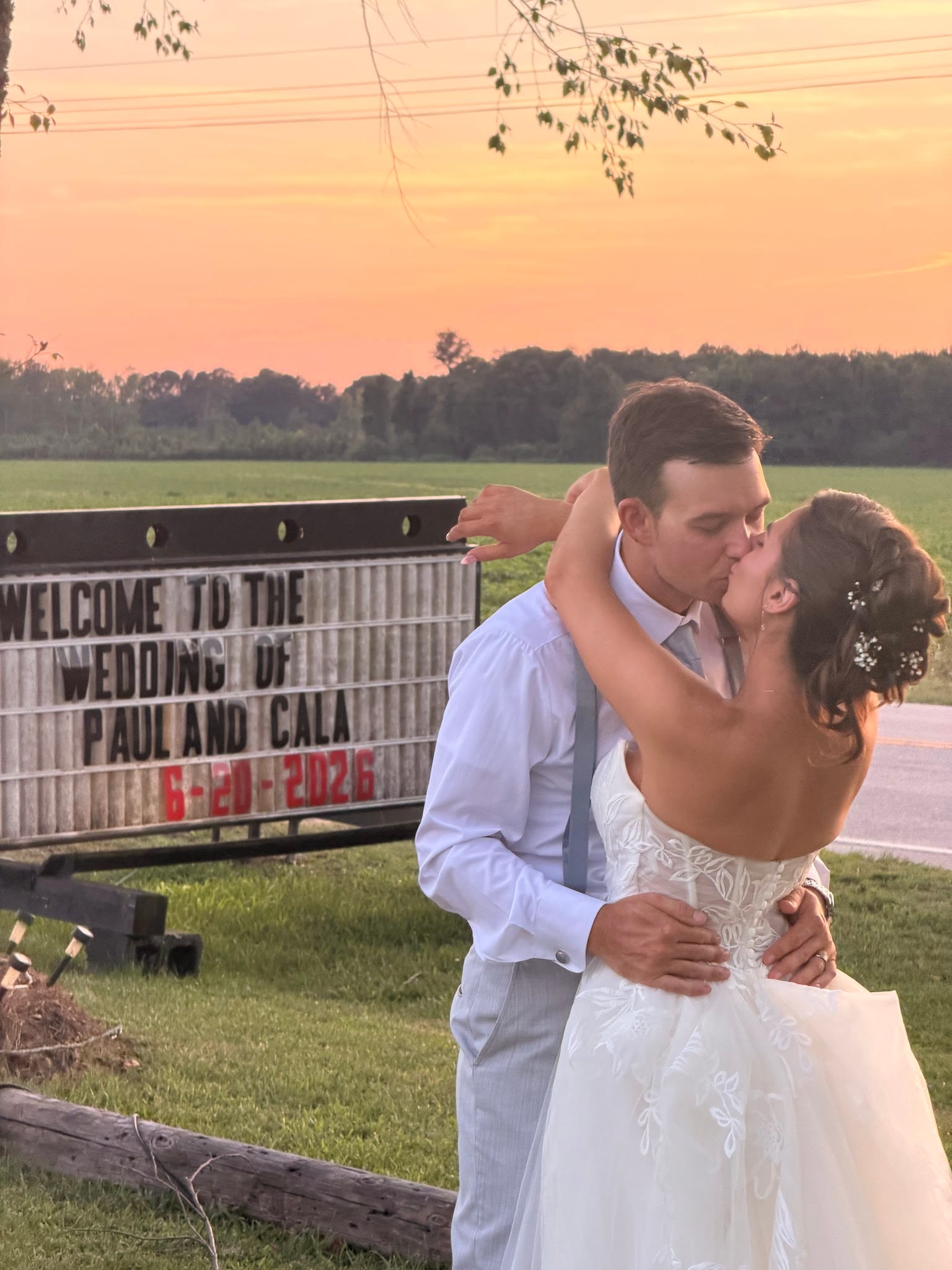 Bride and groom share a kiss in front of the Scarborough House wedding welcome marquee sign at sunset with golden pink skies and green fields in the background.