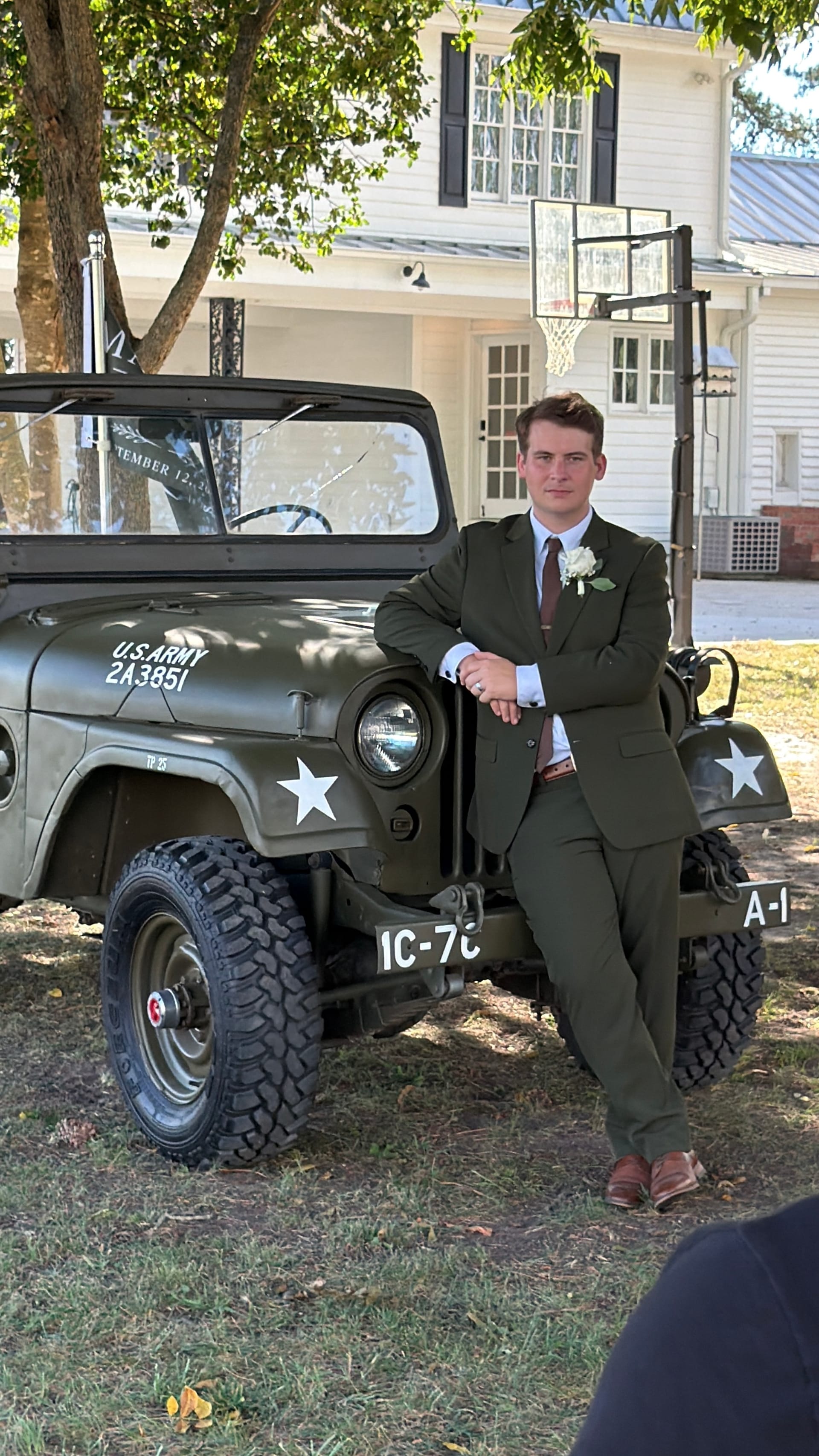 Groom in an olive green suit with a white boutonnière leans casually against a vintage U.S. Army Jeep in front of the historic 1821 Scarborough House manor on a sunny wedding day.