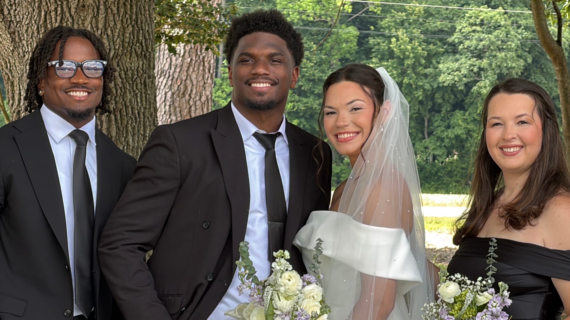 Bride in white off-shoulder gown and veil poses with groom and two wedding guests beneath a large oak tree at Scarborough House estate.