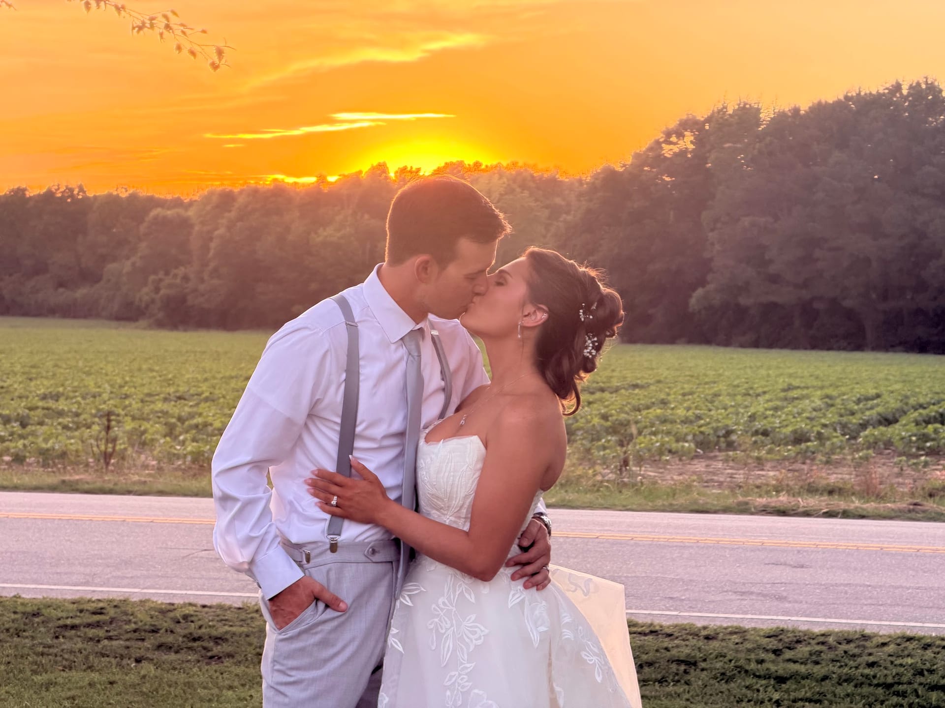 Bride and groom share a romantic kiss in front of a glowing orange sunset over open farmland fields at Scarborough House estate wedding venue.