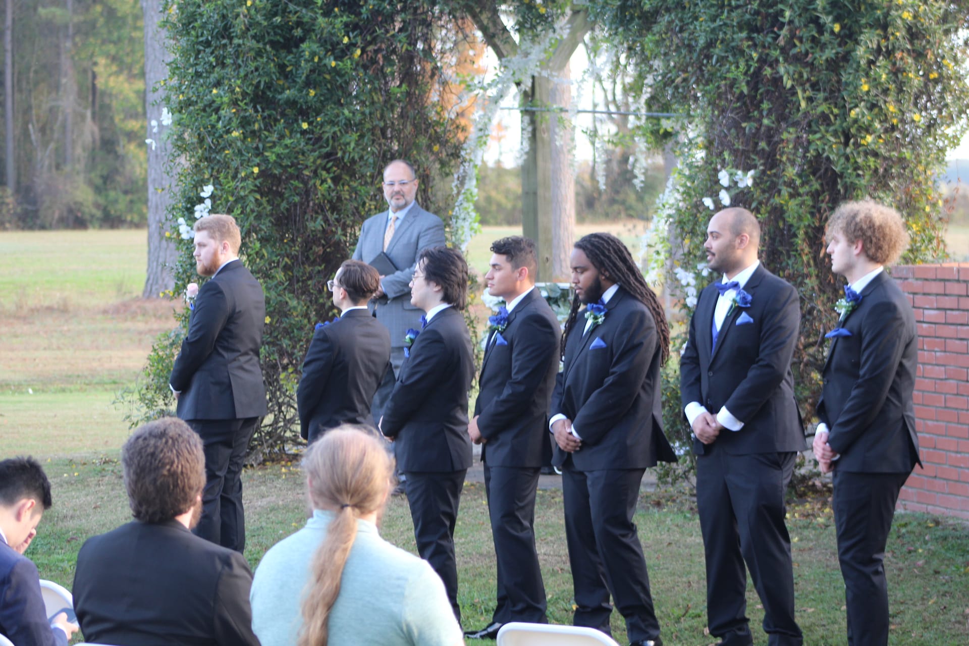 Groomsmen in black tuxedos with blue boutonnieres stand at attention beside a floral arch during an outdoor wedding ceremony at Scarborough House historic estate.