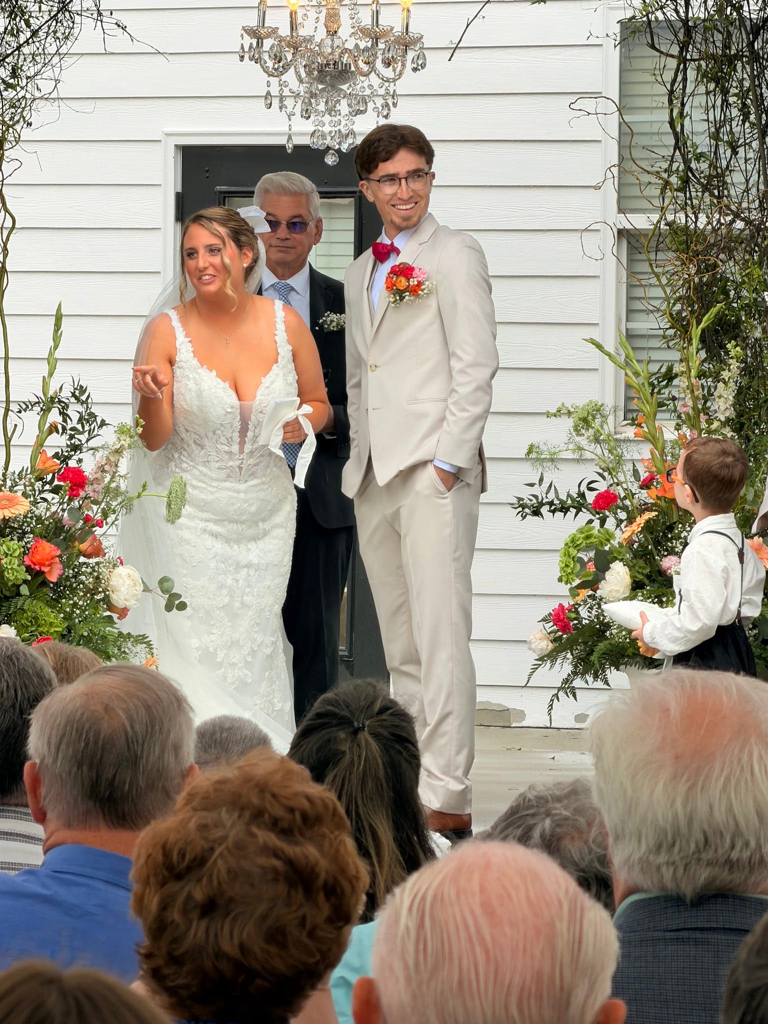 Bride in a lace gown and groom in a tan suit smile joyfully at the altar of Scarborough House, surrounded by vibrant coral and pink floral arrangements against the historic white manor exterior.