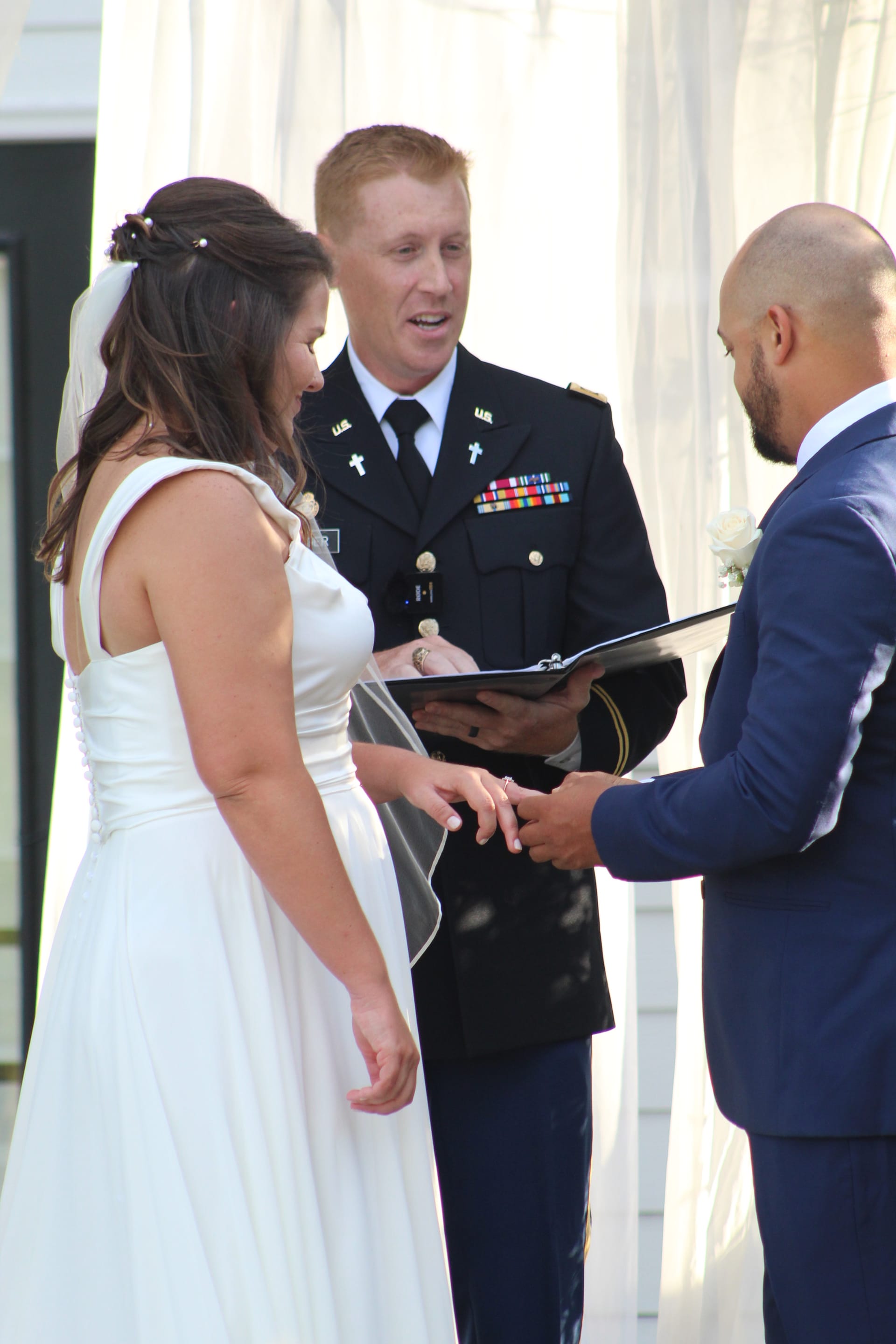A groom in a navy suit places a wedding ring on his brides finger as a military chaplain in dress uniform officiates the outdoor ceremony at Scarborough House.