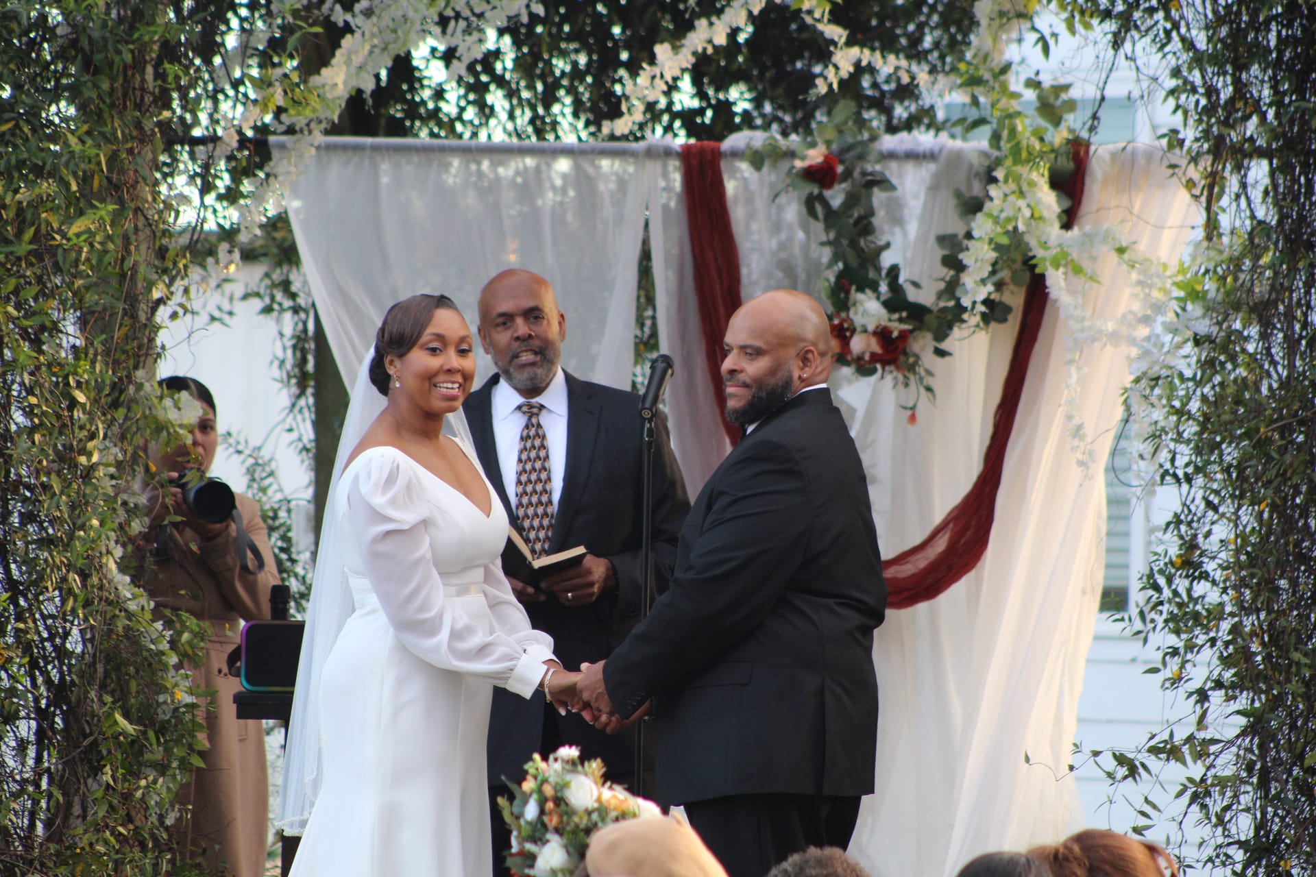 Smiling bride in a long-sleeve white gown and groom in a black tuxedo hold hands during their outdoor wedding ceremony beneath a floral and burgundy-draped arch at Scarborough House historic estate.