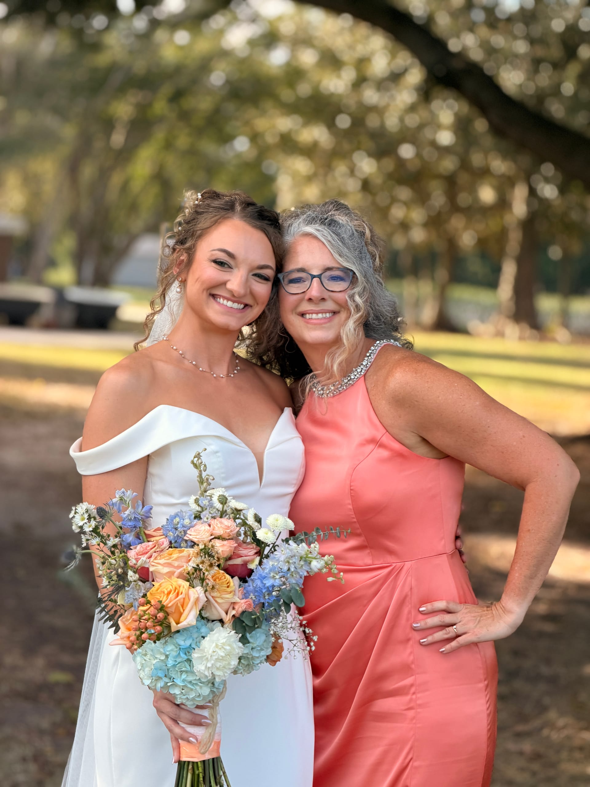 Bride in an off-shoulder white gown holding a pastel bouquet poses cheek-to-cheek with a woman in a coral dress beneath the sprawling oak trees at Scarborough House.
