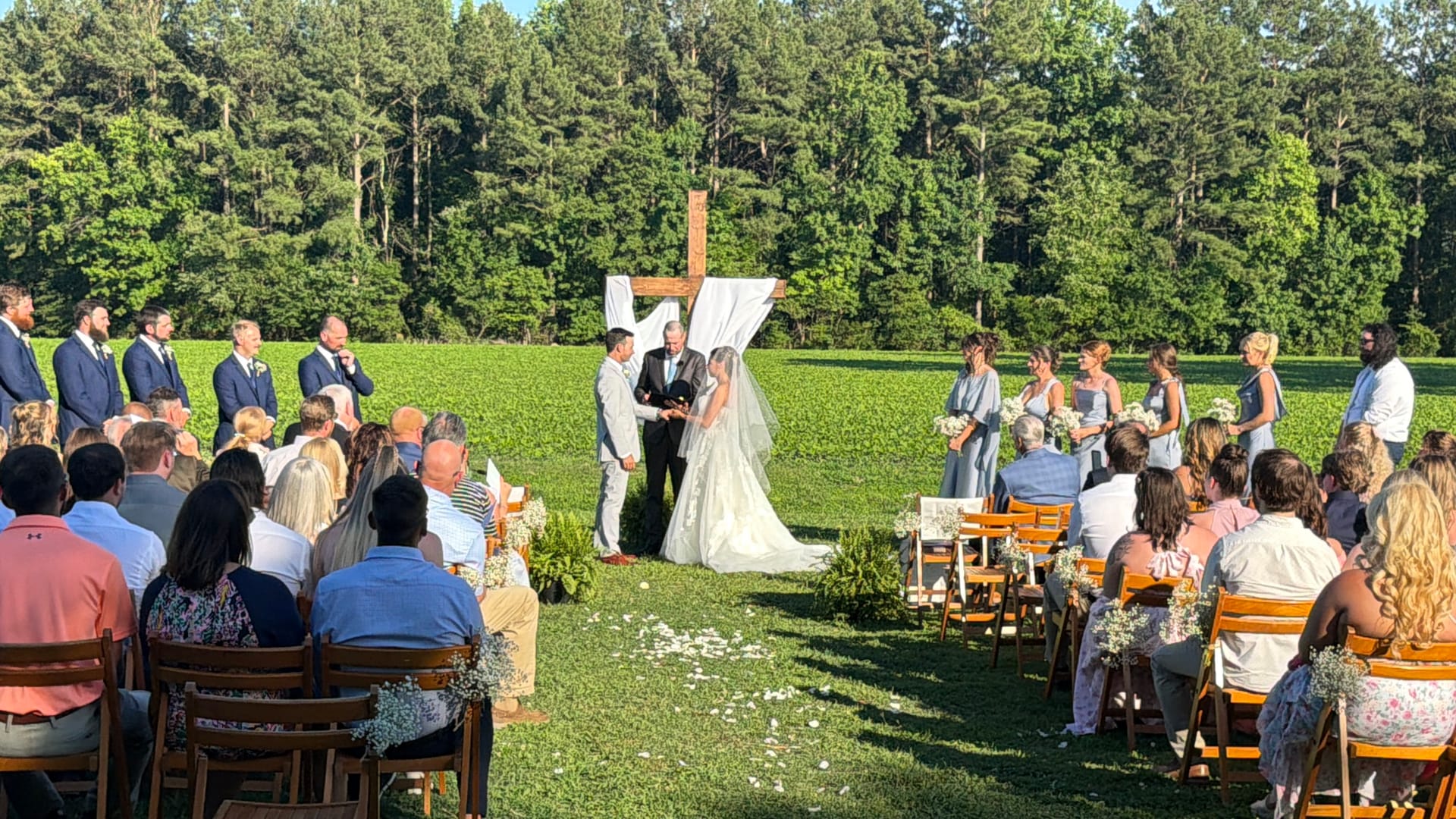 Outdoor wedding ceremony on a sunlit lawn at Scarborough House with the couple exchanging vows beneath a draped wooden cross, surrounded by their wedding party and seated guests with a green field and tall trees in the background.