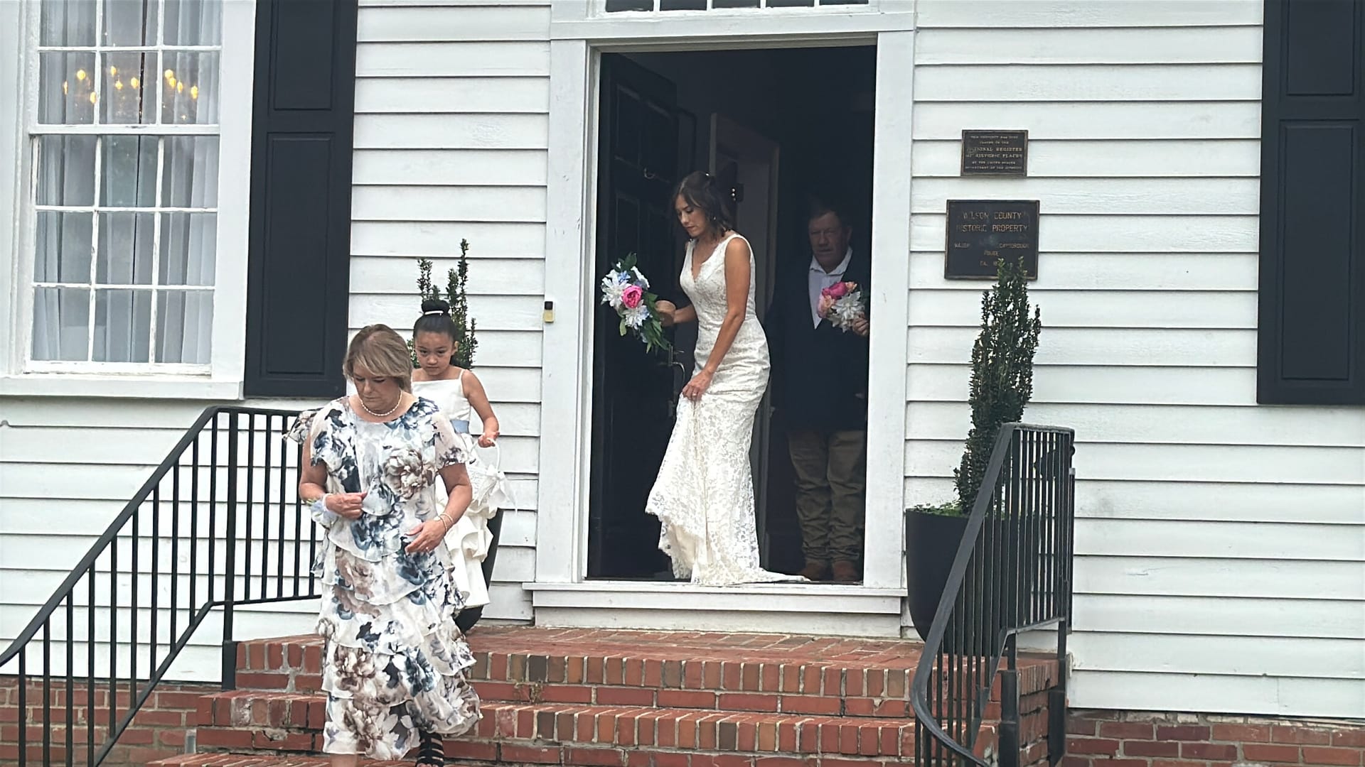 Bride in a lace gown holding a pink and white bouquet steps out of the historic 1821 Scarborough House manor door accompanied by a flower girl and guests on brick front steps.