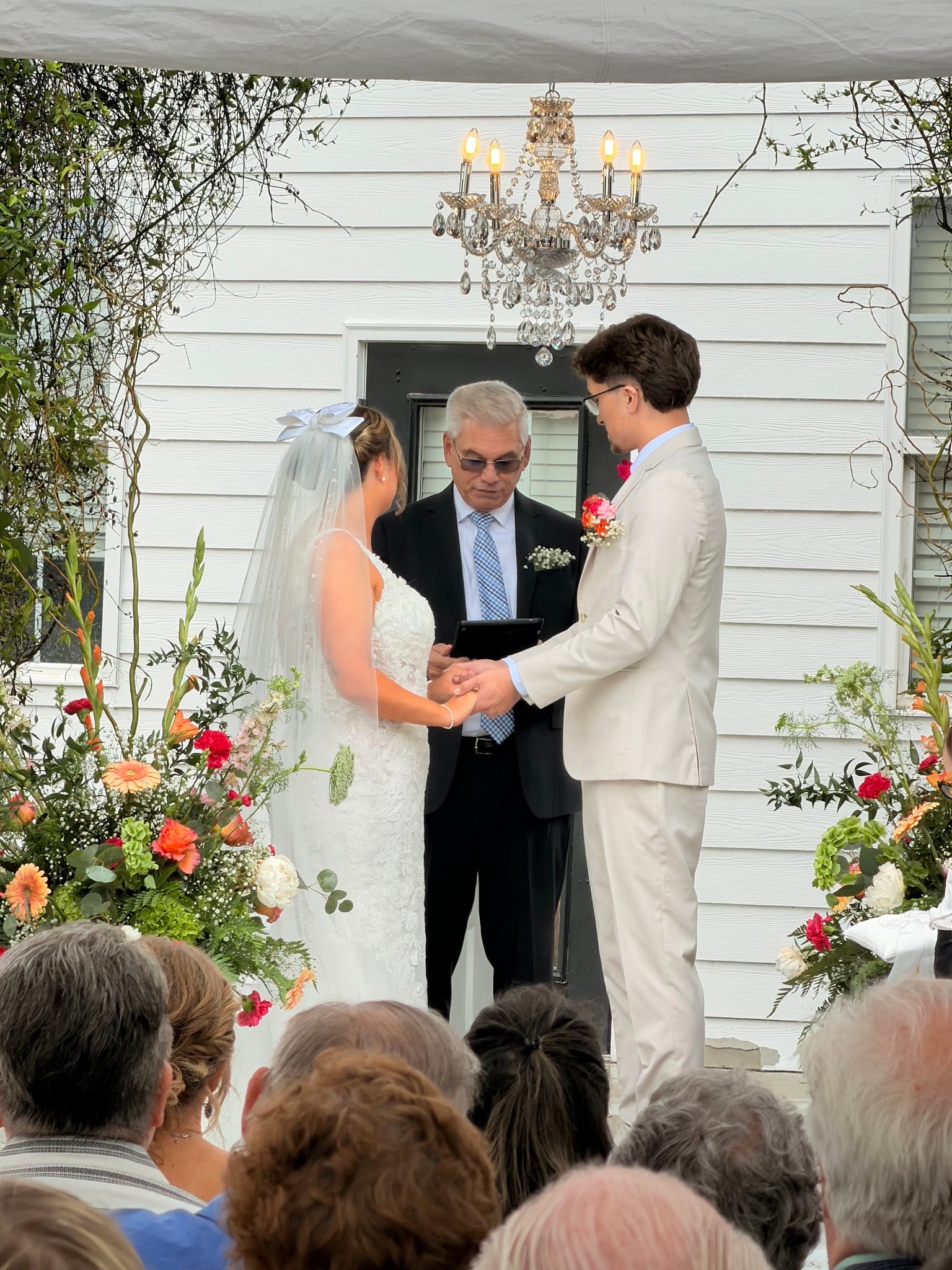 Bride and groom exchange rings during an outdoor ceremony at a historic white manor estate, framed by vibrant orange and pink floral arrangements and an elegant crystal chandelier.