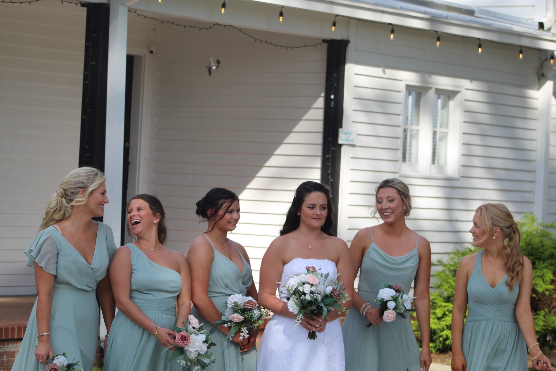 Bride in white gown stands with five bridesmaids in sage green dresses holding floral bouquets in front of the historic white clapboard facade of Scarborough House.