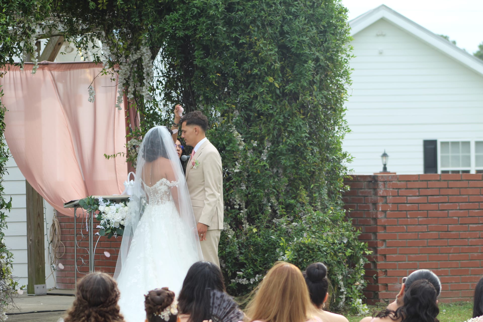 Bride in a lace ball gown with veil and groom in a tan suit hold hands at an outdoor altar adorned with white florals and blush pink draping at Scarborough House.