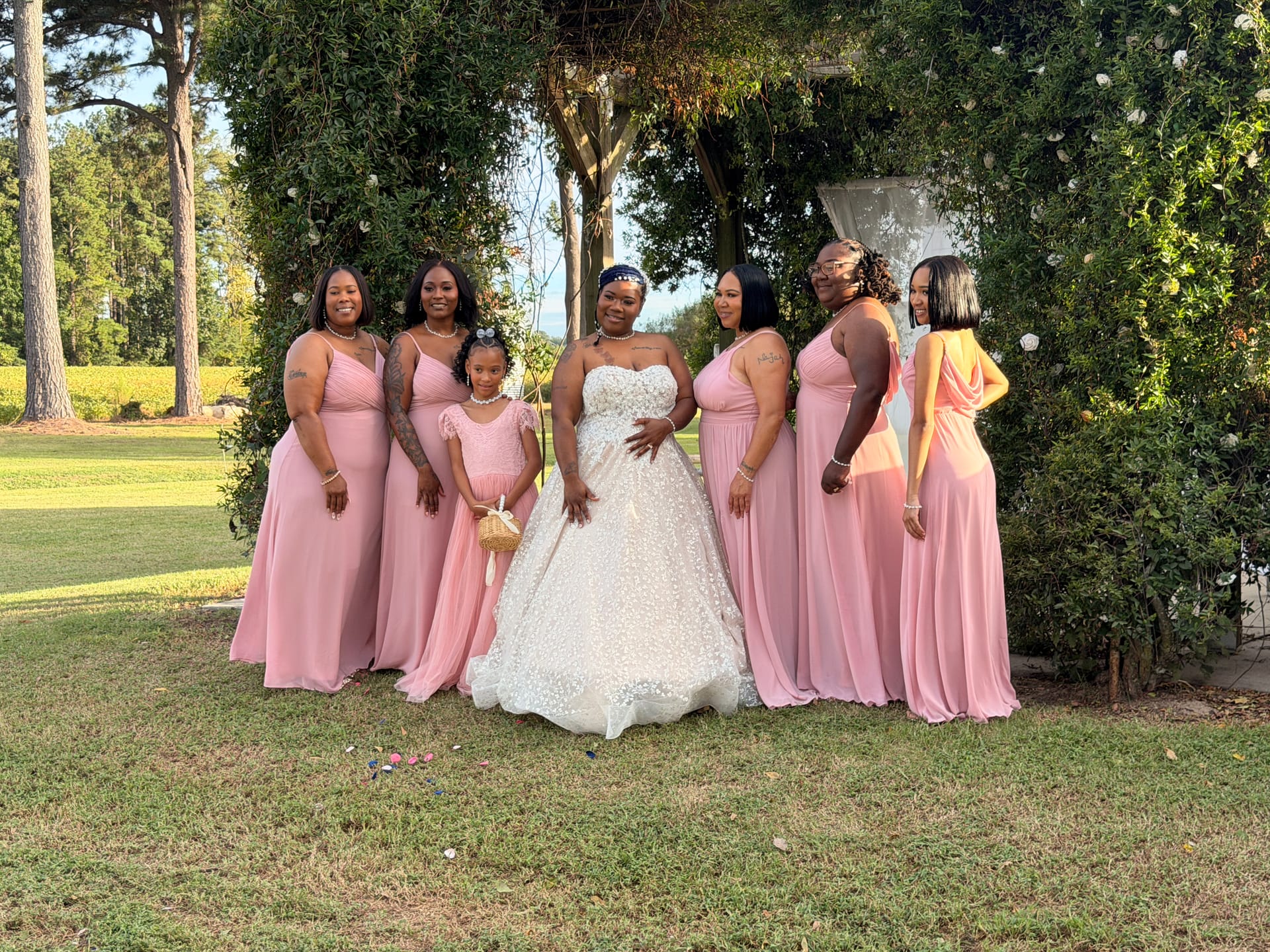 Bride in a floral ballgown poses with five bridesmaids in blush pink gowns and a flower girl beneath a vine-covered arch on the ceremony lawn at Scarborough House.