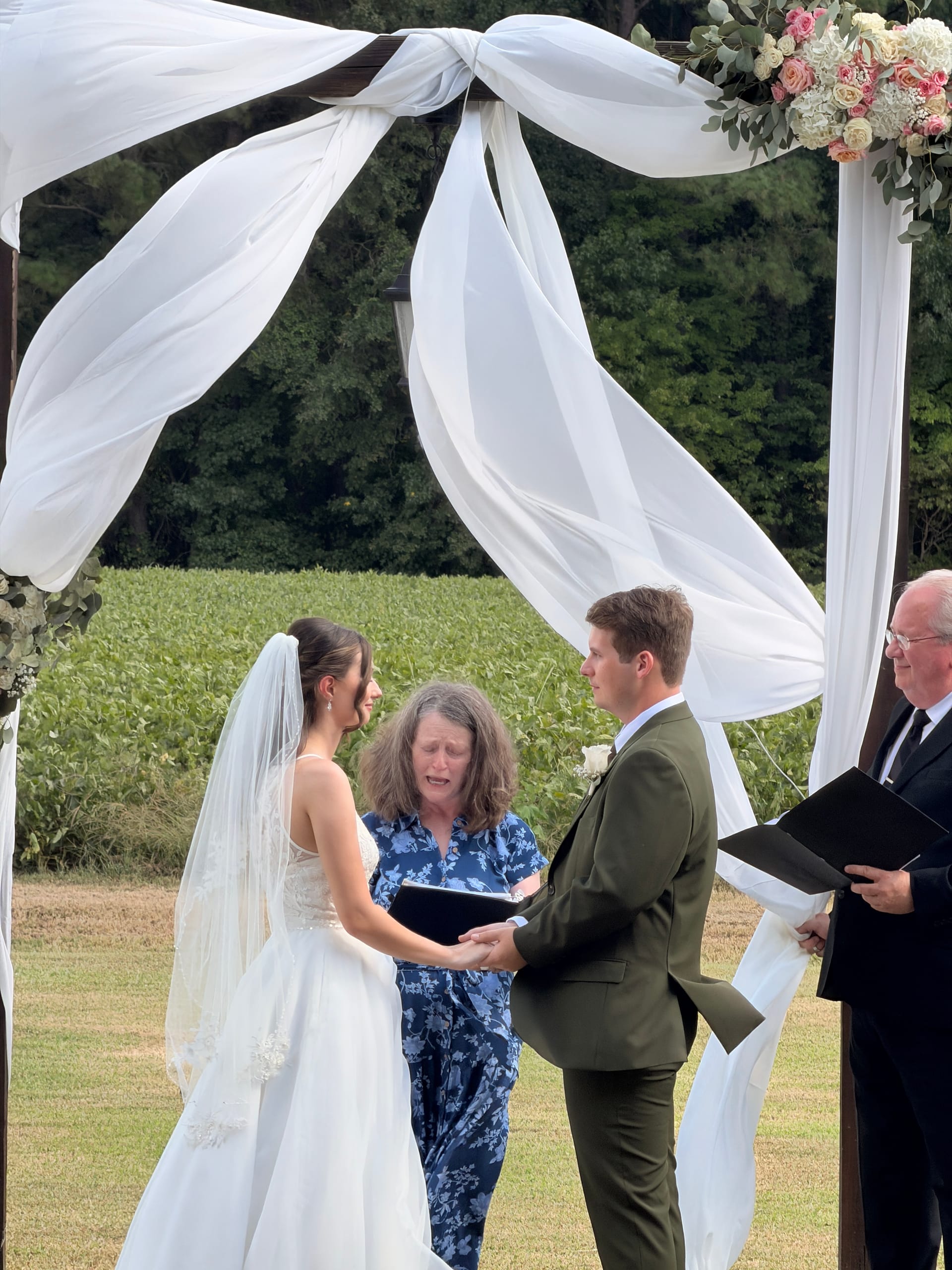 Bride and groom exchange vows beneath a white draped wooden arch adorned with pink and ivory florals on the ceremony lawn at Scarborough House, with lush green farmland fields in the background.
