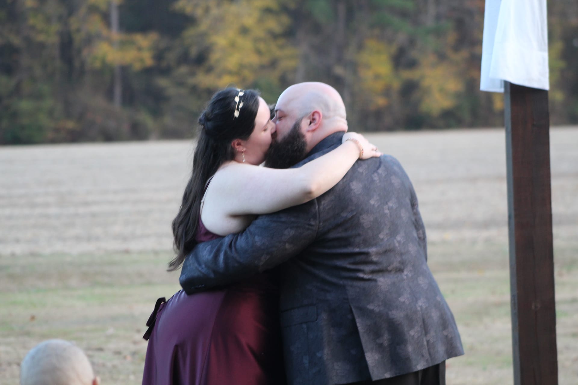 Bride in burgundy gown and groom in a patterned suit share their first kiss during an outdoor autumn ceremony on the Scarborough House estate lawn with golden fall trees in the background.