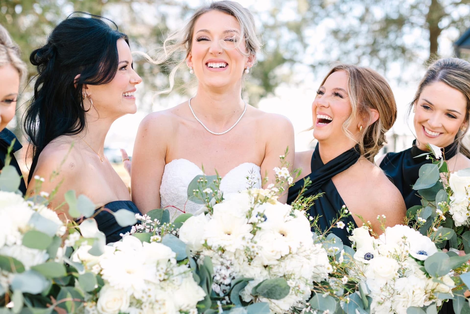 Bride in a strapless white gown laughing joyfully with her bridesmaids in black dresses holding lush white floral bouquets at Scarborough House.