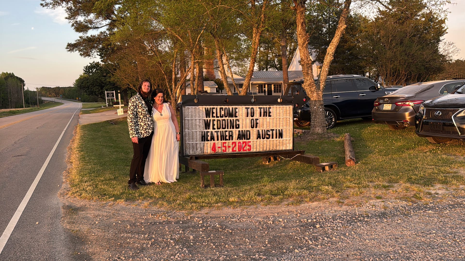 Bride in a white gown and groom in a floral blazer pose together beside a marquee sign welcoming guests to the wedding of Heather and Austin on April 5 2025 at Scarborough House during golden hour.