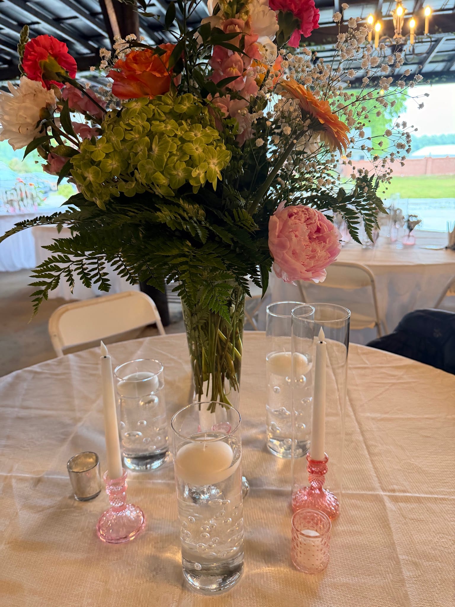 Elegant wedding table centerpiece featuring a tall vase of pink peonies, green hydrangeas, and mixed florals surrounded by floating candle cylinder vases and pink glass bud vases on a white linen tablecloth inside Scarborough House.