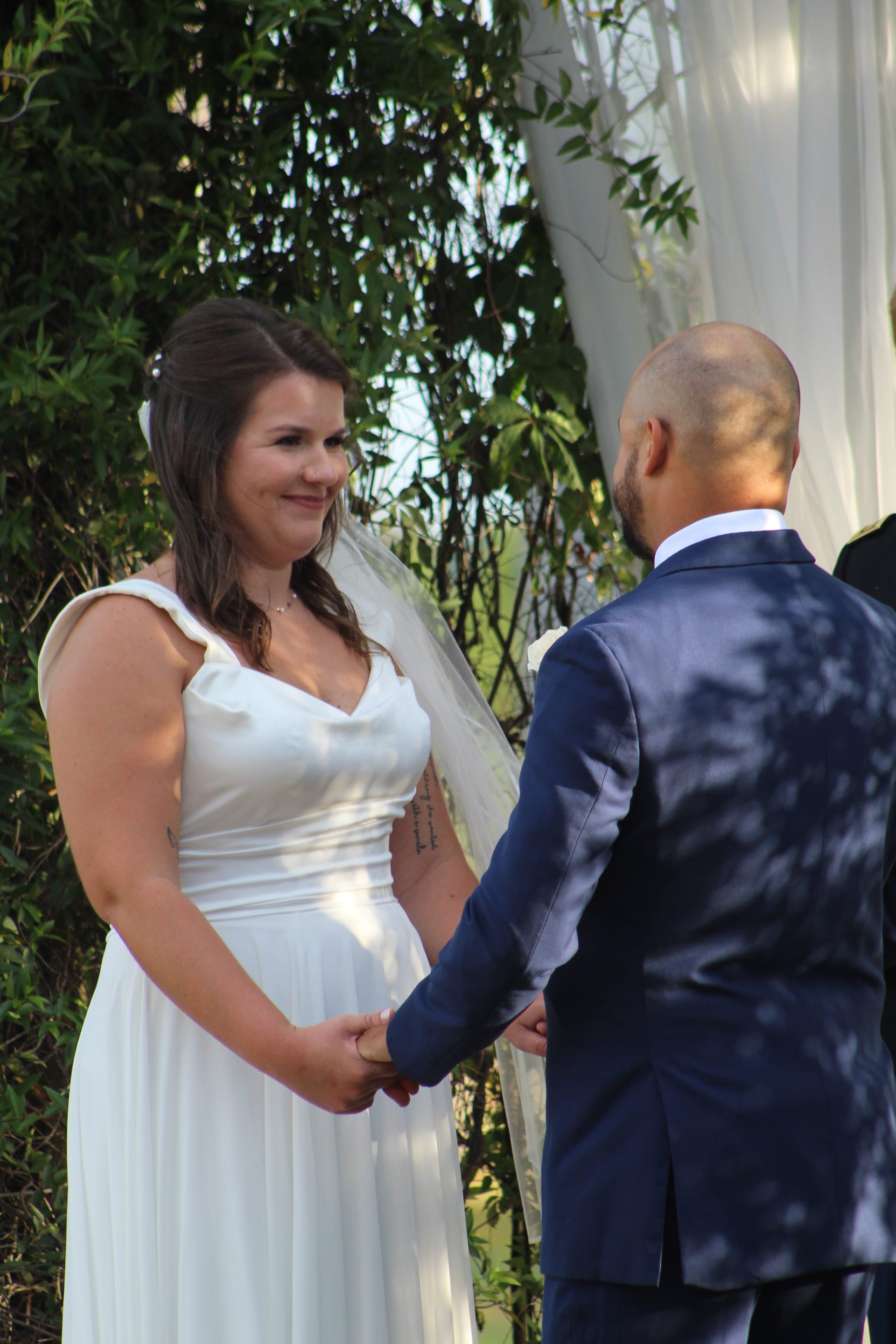 Bride in a flowing white gown smiles warmly at her groom while holding hands during an outdoor wedding ceremony framed by lush greenery and a white draped arch at Scarborough House.