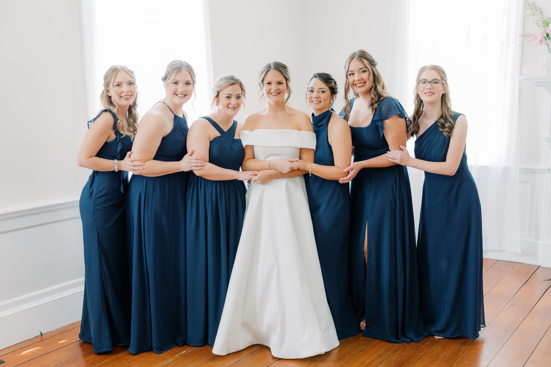 Bride in an off-shoulder white gown surrounded by six bridesmaids in navy blue dresses posing together in a bright white interior room at Scarborough House.