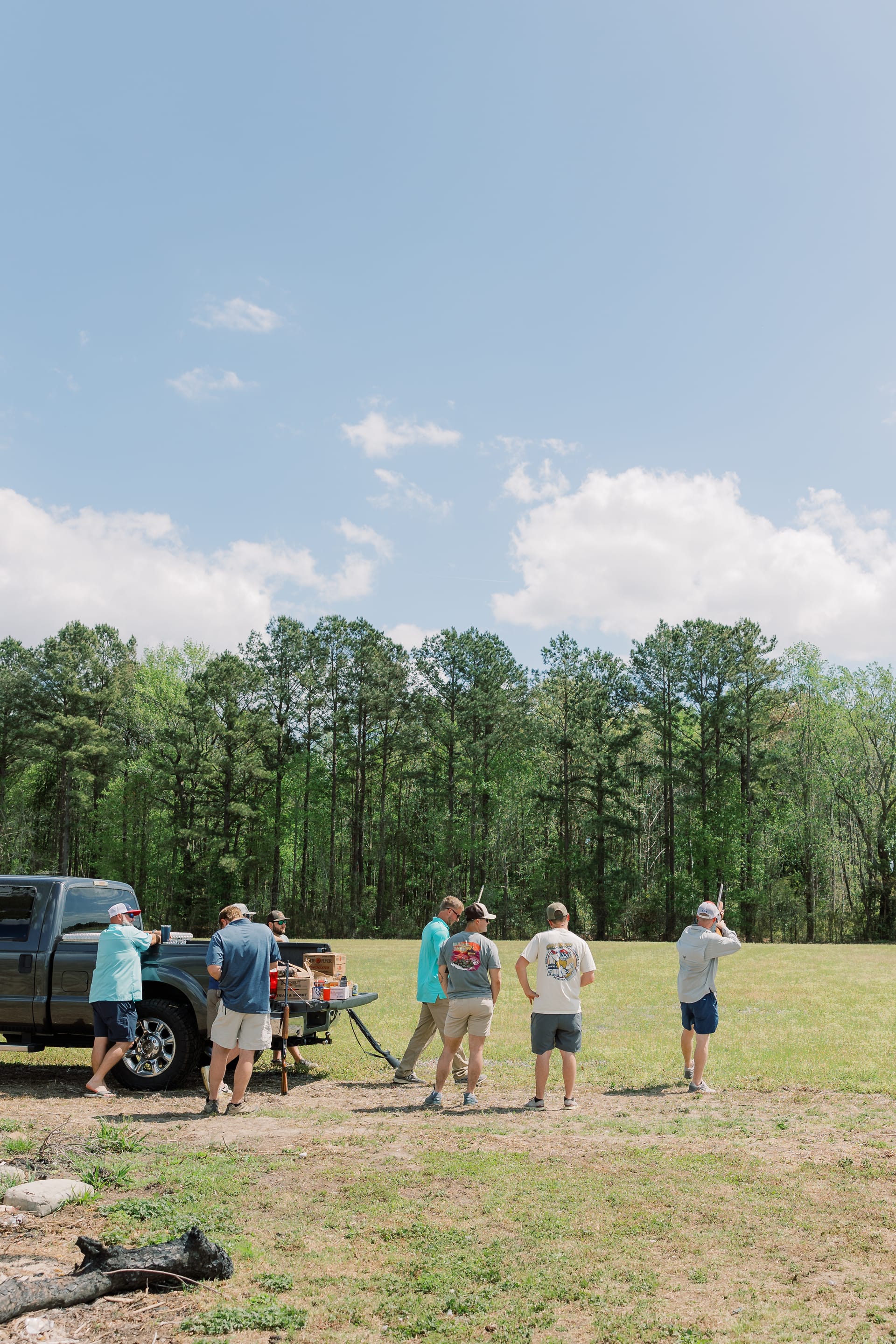 A group of groomsmen shoot clay targets in an open field beside a pickup truck at Scarborough House with pine trees in the background.