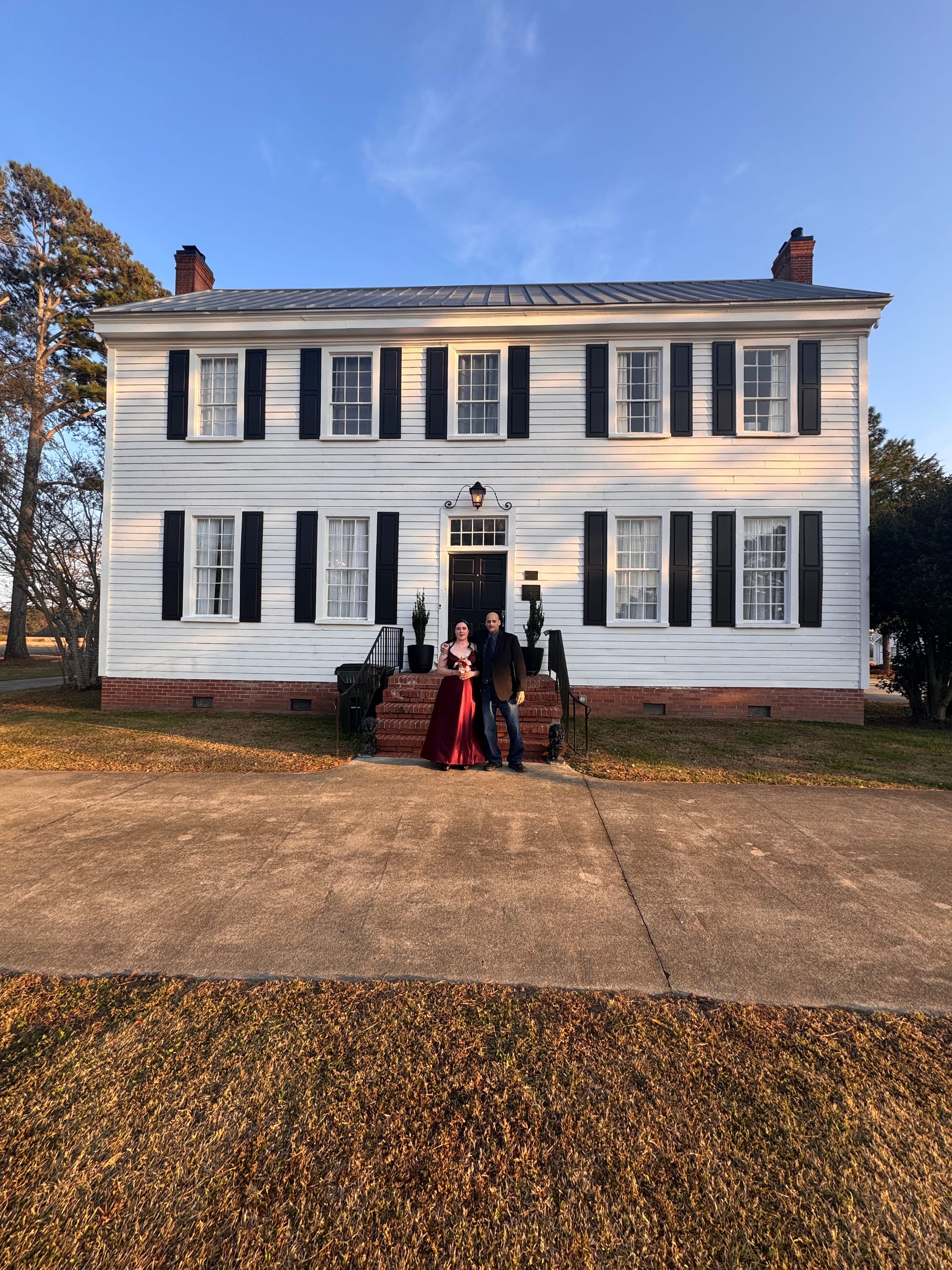 A couple in formal attire poses on the front steps of the 1821 Scarborough House white colonial manor with black shutters at golden hour.