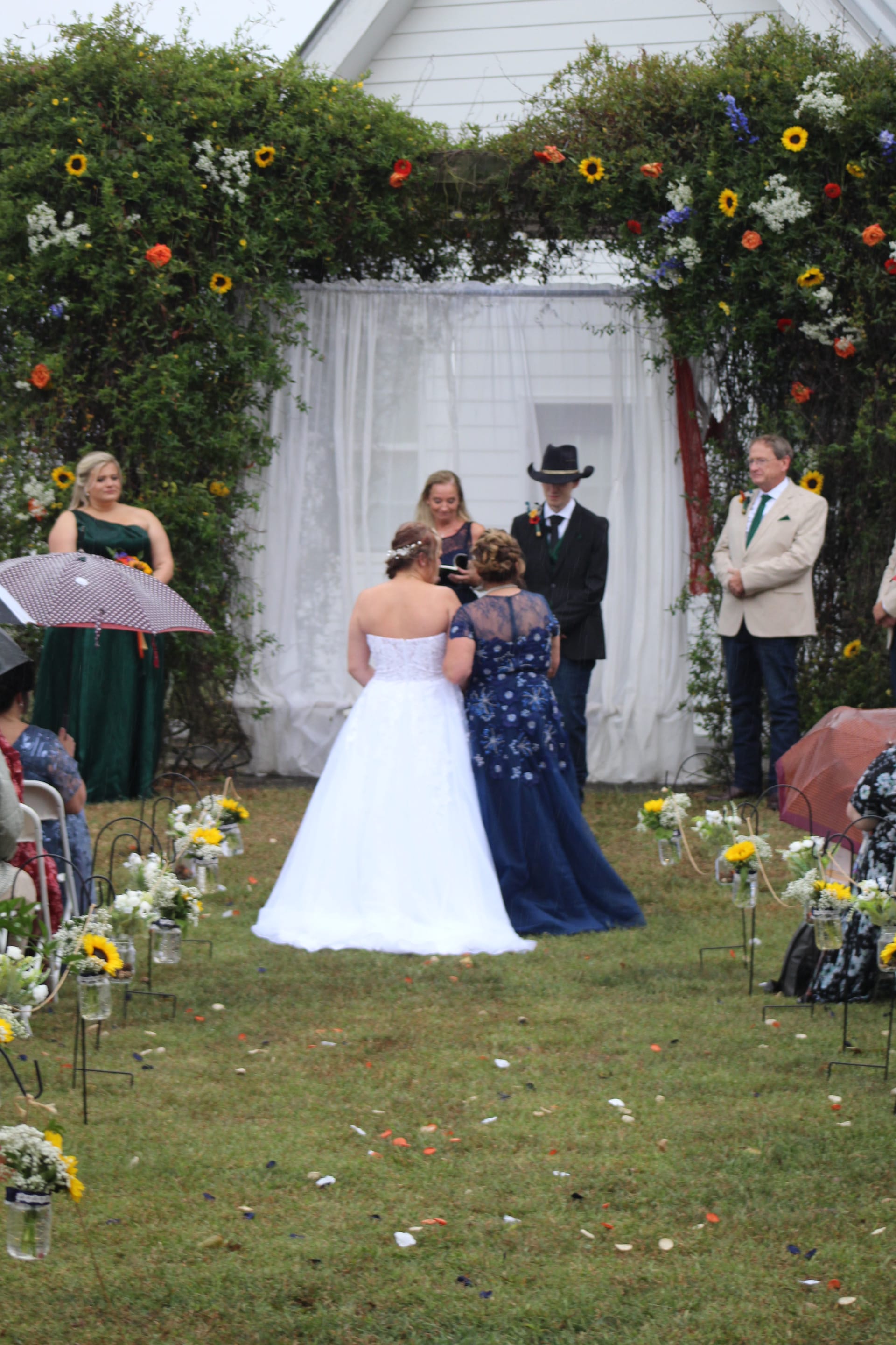Outdoor wedding ceremony on the lawn beneath a lush floral arch decorated with sunflowers and wildflowers as the bride in white and a guest in navy blue stand before the officiant and groom in a cowboy hat.