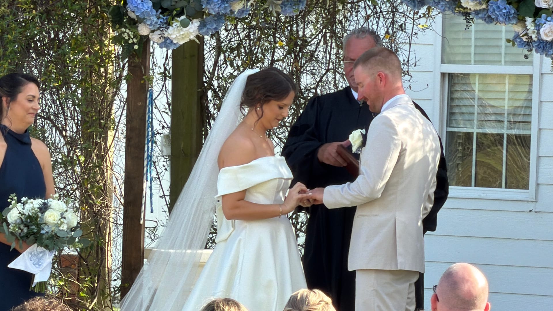 Groom placing a wedding ring on the bride's finger beneath a blue hydrangea floral arch at an outdoor ceremony while the officiant looks on.