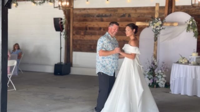 Bride in an elegant off-shoulder white gown shares a tender dance with her father inside the rustic grand hall at Scarborough House, with floral decor and wedding cake table visible in the background.