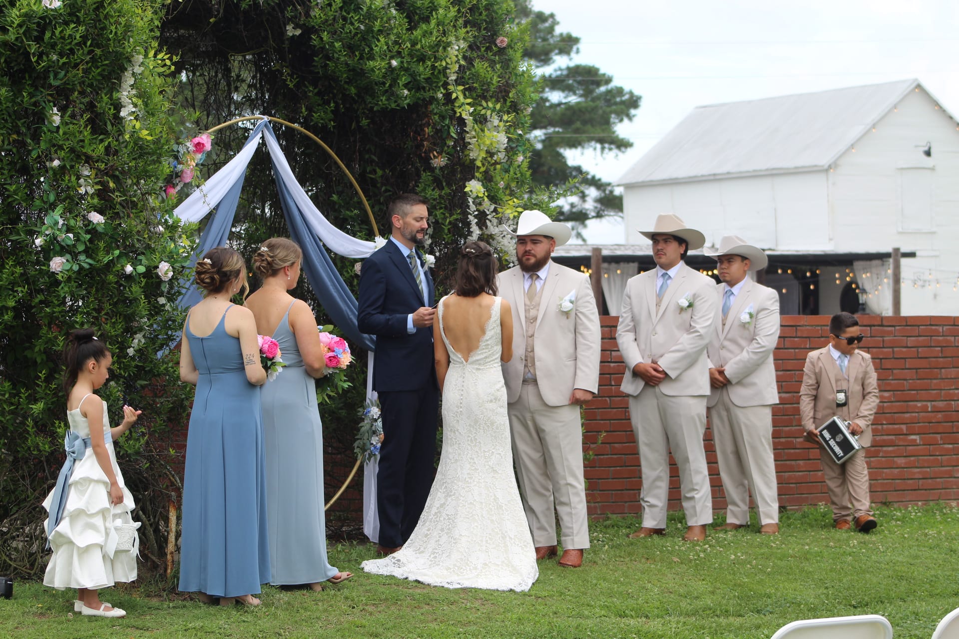 A bride and groom exchange vows beneath a floral hoop arch with blue draping on the ceremony lawn, surrounded by bridesmaids in dusty blue gowns and groomsmen in tan suits and cowboy hats, with a white barn visible in the background.