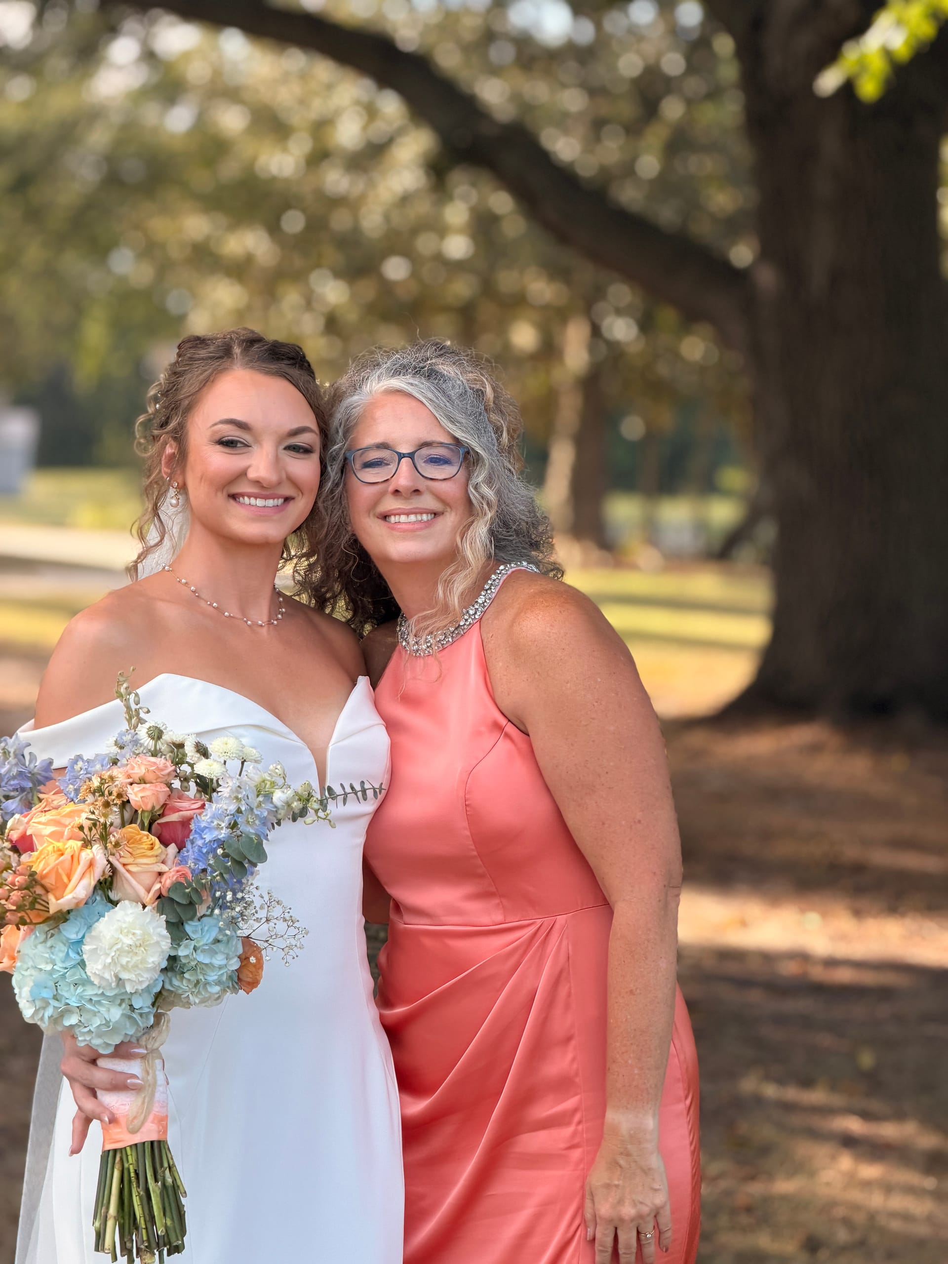 Bride in a white off-shoulder gown holding a pastel peach and blue bouquet poses with an older woman in a coral dress beneath the grand oak trees at Scarborough House.