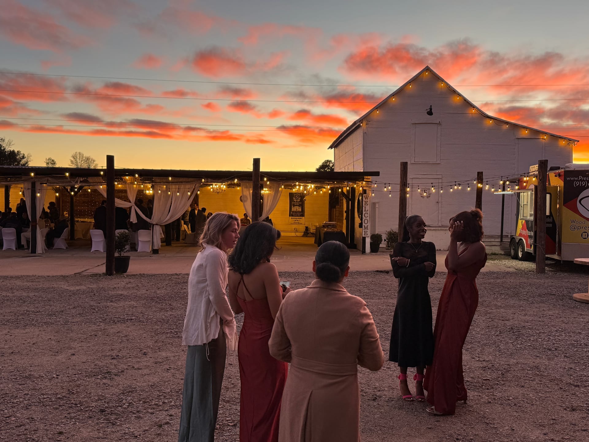 Wedding guests mingle in a gravel courtyard at sunset beside a lit white barn.