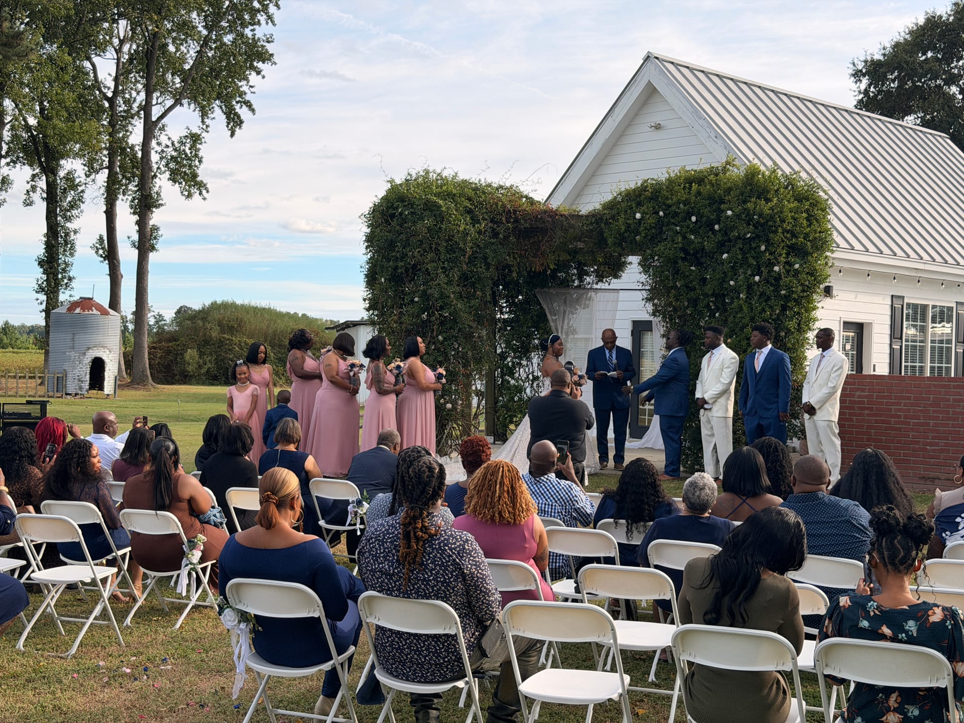 Wedding ceremony on the lawn at Scarborough House with bridesmaids in dusty rose gowns and groomsmen in navy suits standing beneath a lush greenery arch in front of the white historic barn.