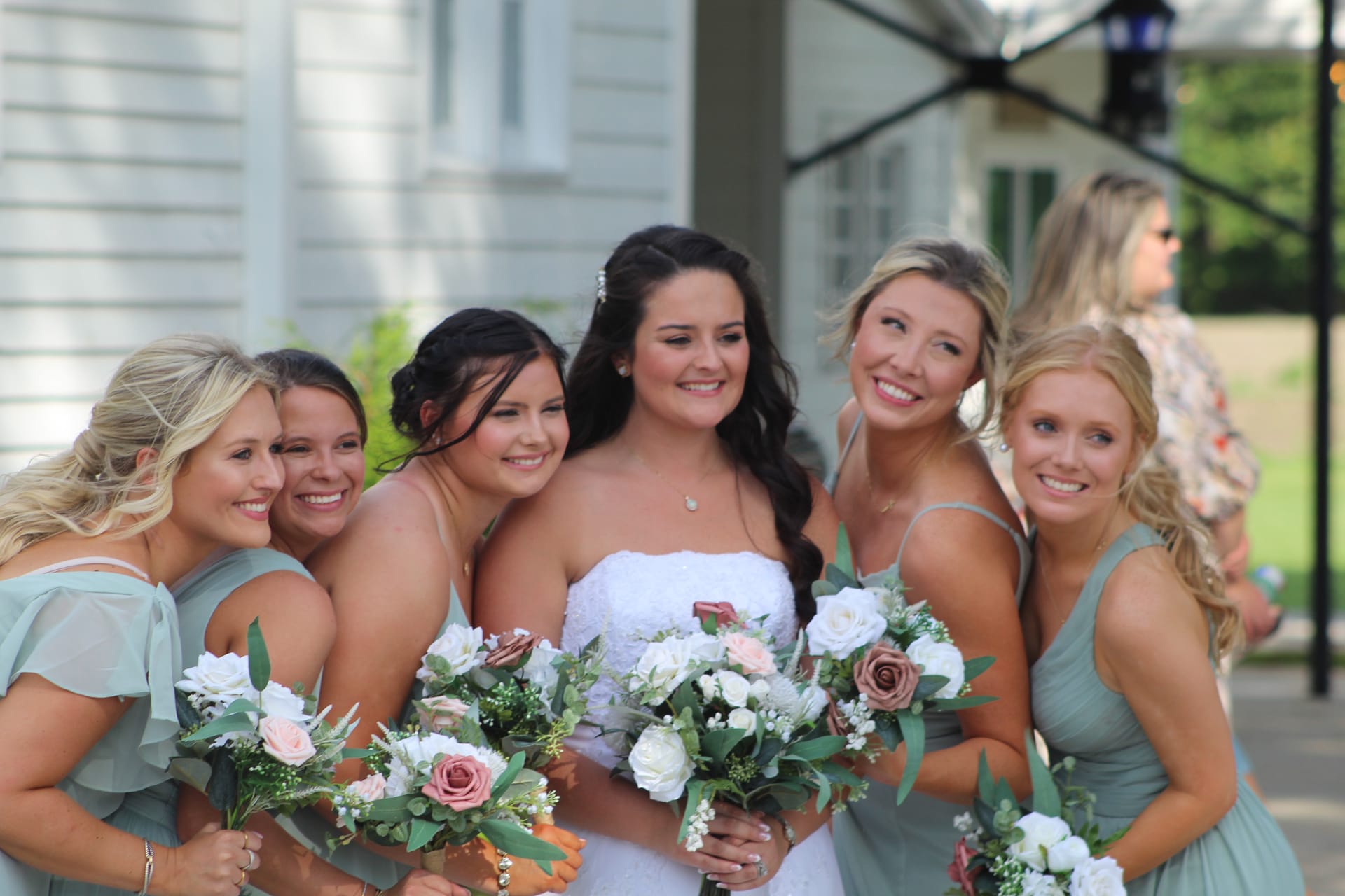 Smiling bride in a white strapless gown poses with four bridesmaids in sage green dresses holding blush and white floral bouquets outside the historic Scarborough House estate.