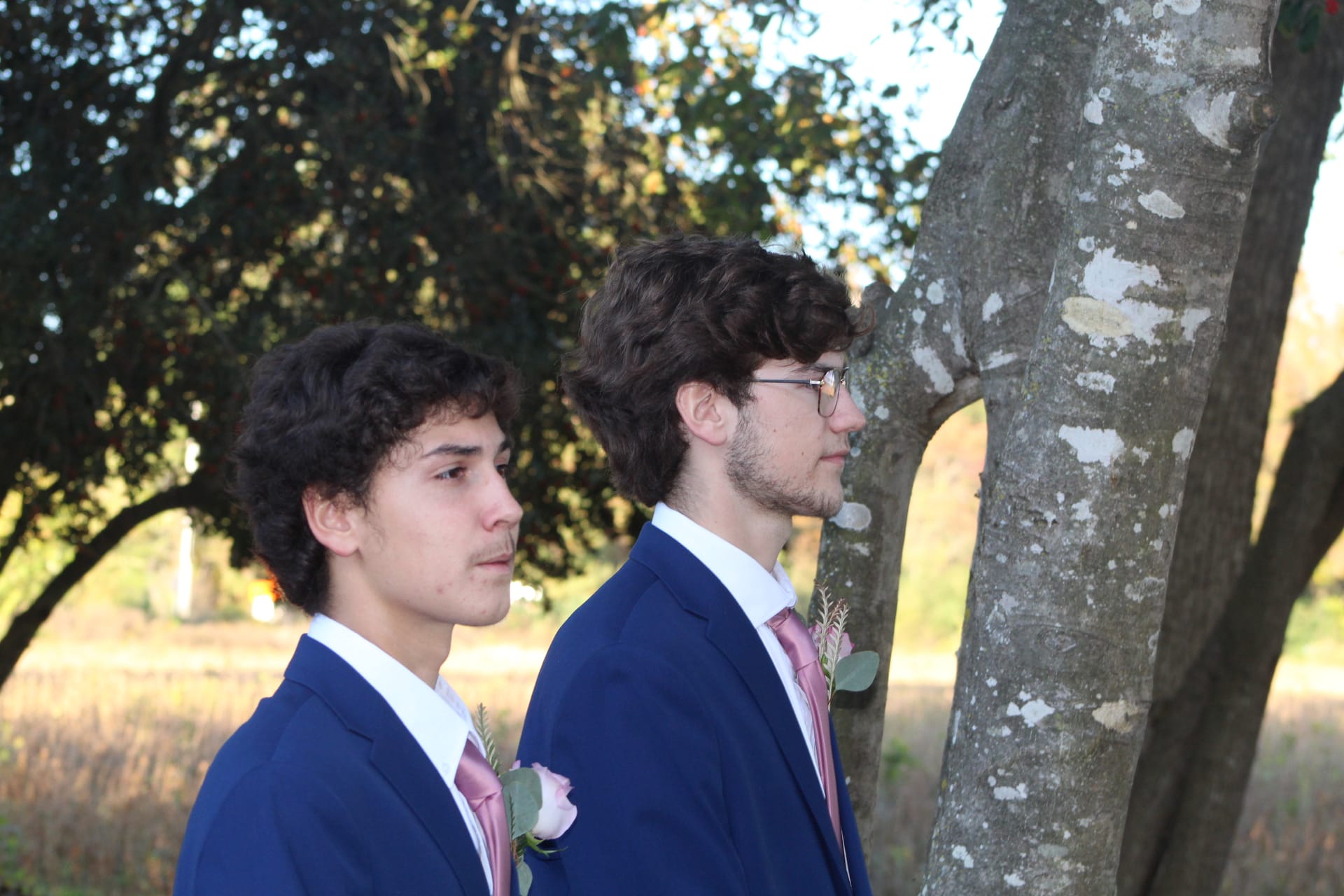 Two groomsmen in navy suits with blush pink ties and boutonnieres standing in profile among the oak trees at Scarborough House with open farmland fields in the background.