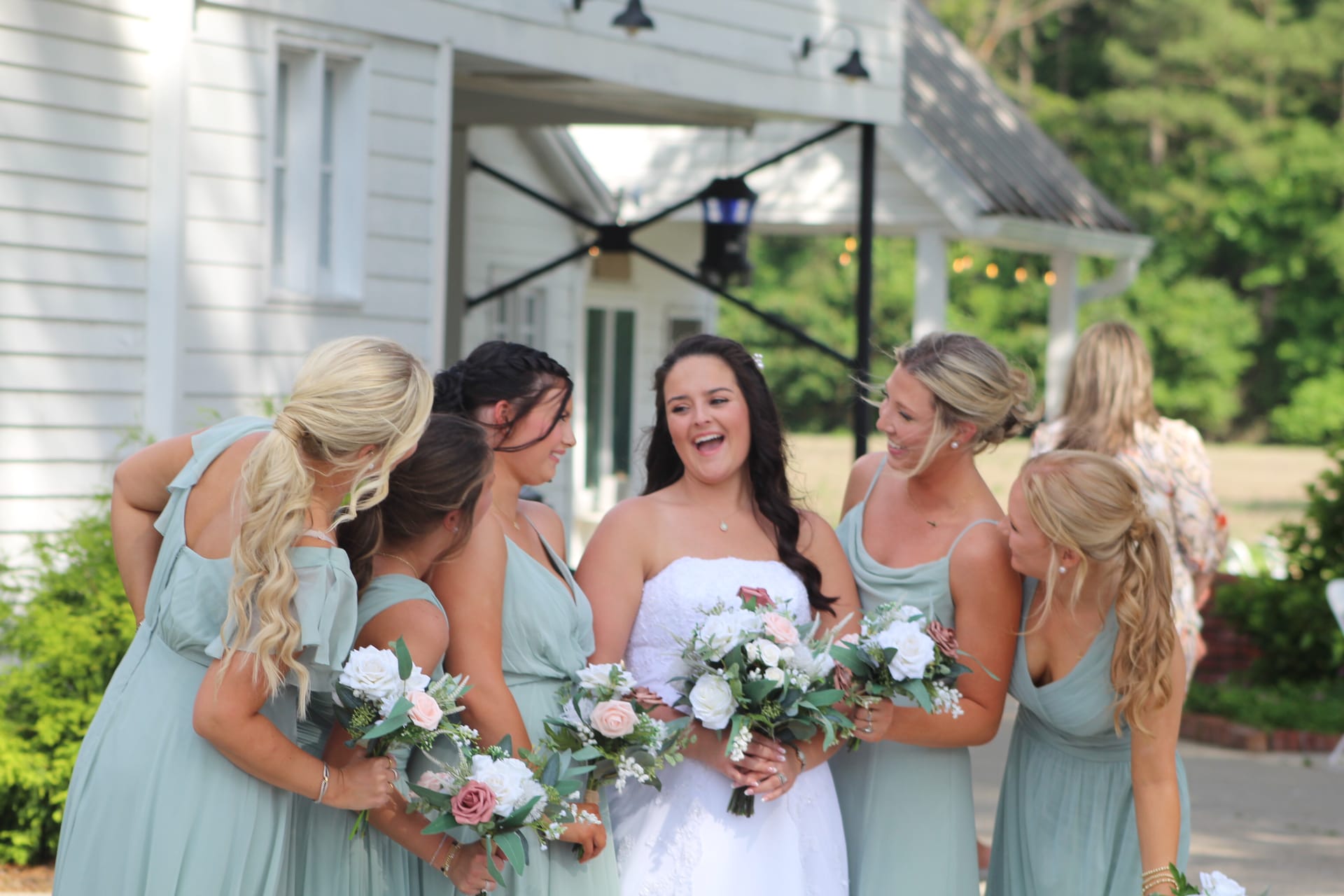 A laughing bride in a white strapless gown holds a white and blush bouquet while her bridesmaids in sage green dresses gather around her in front of the historic Scarborough House manor.