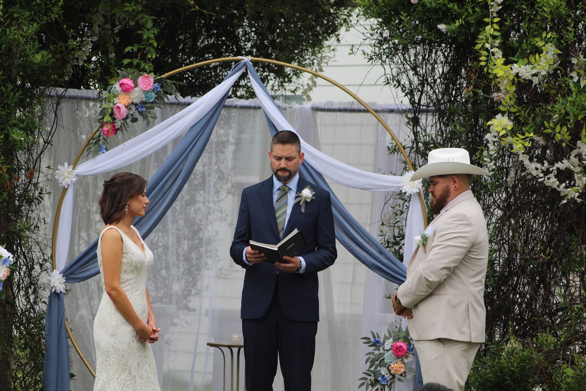 Bride in white lace gown and groom in cream suit with cowboy hat stand before a gold circular arch draped in dusty blue fabric while the officiant reads during an outdoor wedding ceremony at Scarborough House.