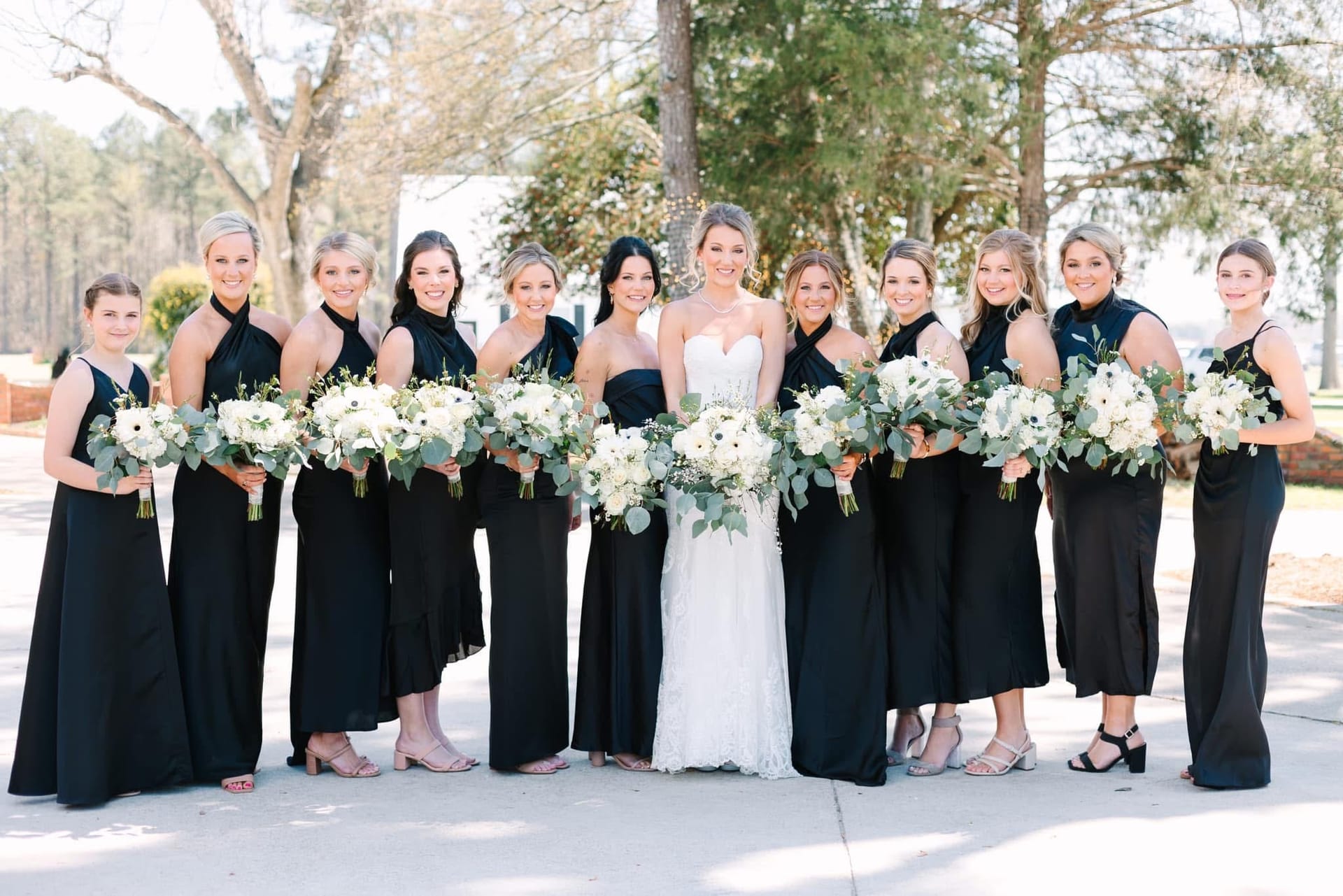 Bride in a white lace gown stands at center surrounded by eleven bridesmaids in black gowns holding white floral bouquets with eucalyptus greenery on the grounds of Scarborough House.