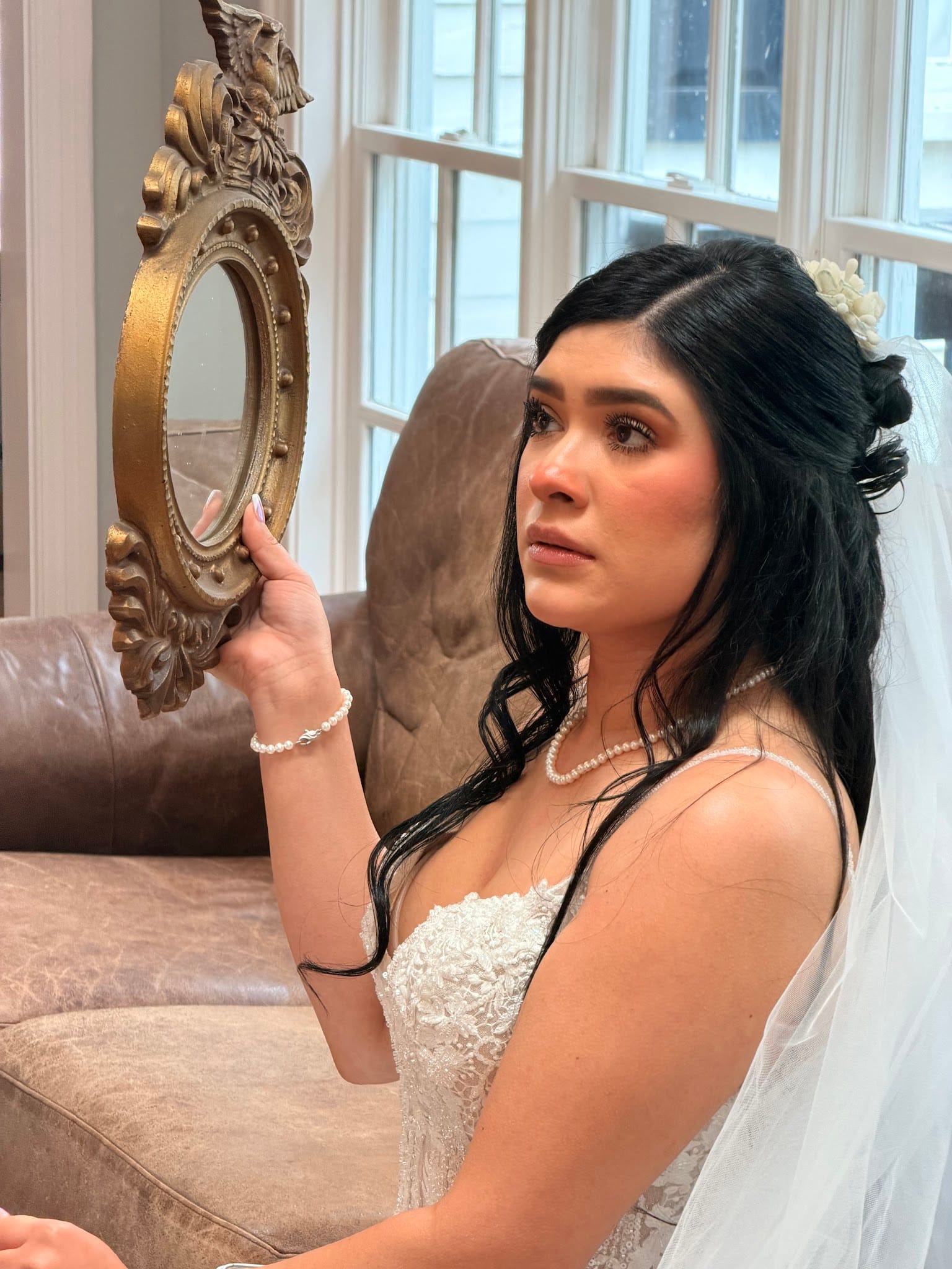 Bride in a lace gown and veil holds an ornate gold hand mirror while seated on a leather sofa near a sunlit window at Scarborough House.