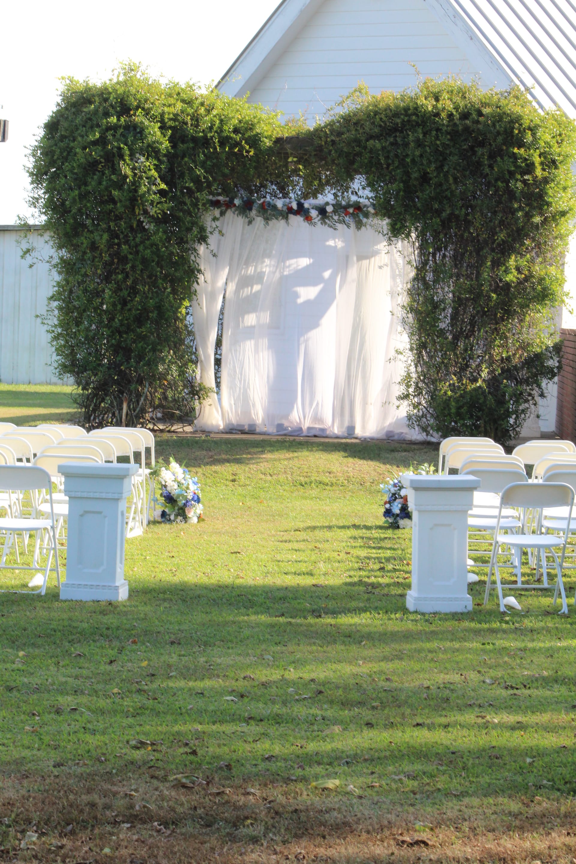 Outdoor wedding ceremony setup at Scarborough House featuring a lush green hedge arch draped in white fabric with red and blue floral accents, flanked by rows of white folding chairs and white pillar pedestals on a manicured lawn.