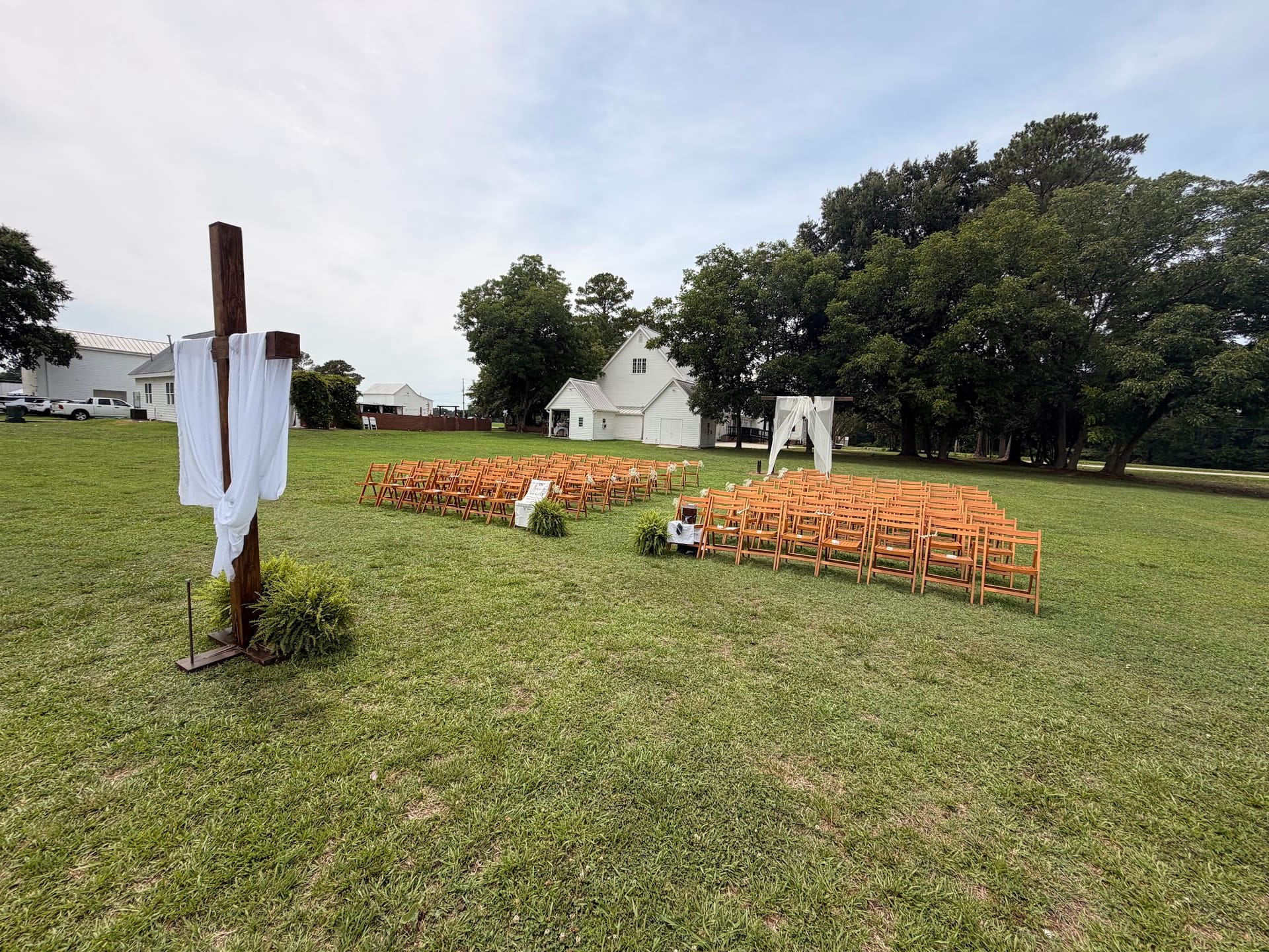 Outdoor wedding ceremony setup at Scarborough House featuring a wooden cross draped in white fabric, rows of wooden folding chairs, a white fabric arbor, and the historic white manor house framed by large oak trees in the background.